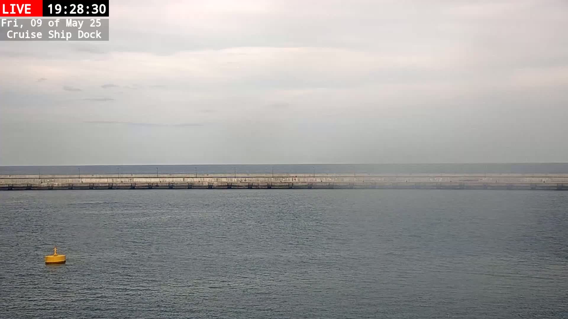 A long concrete wall stretches across a calm, gray-sky day over a body of water, with a single yellow buoy visible in the foreground.