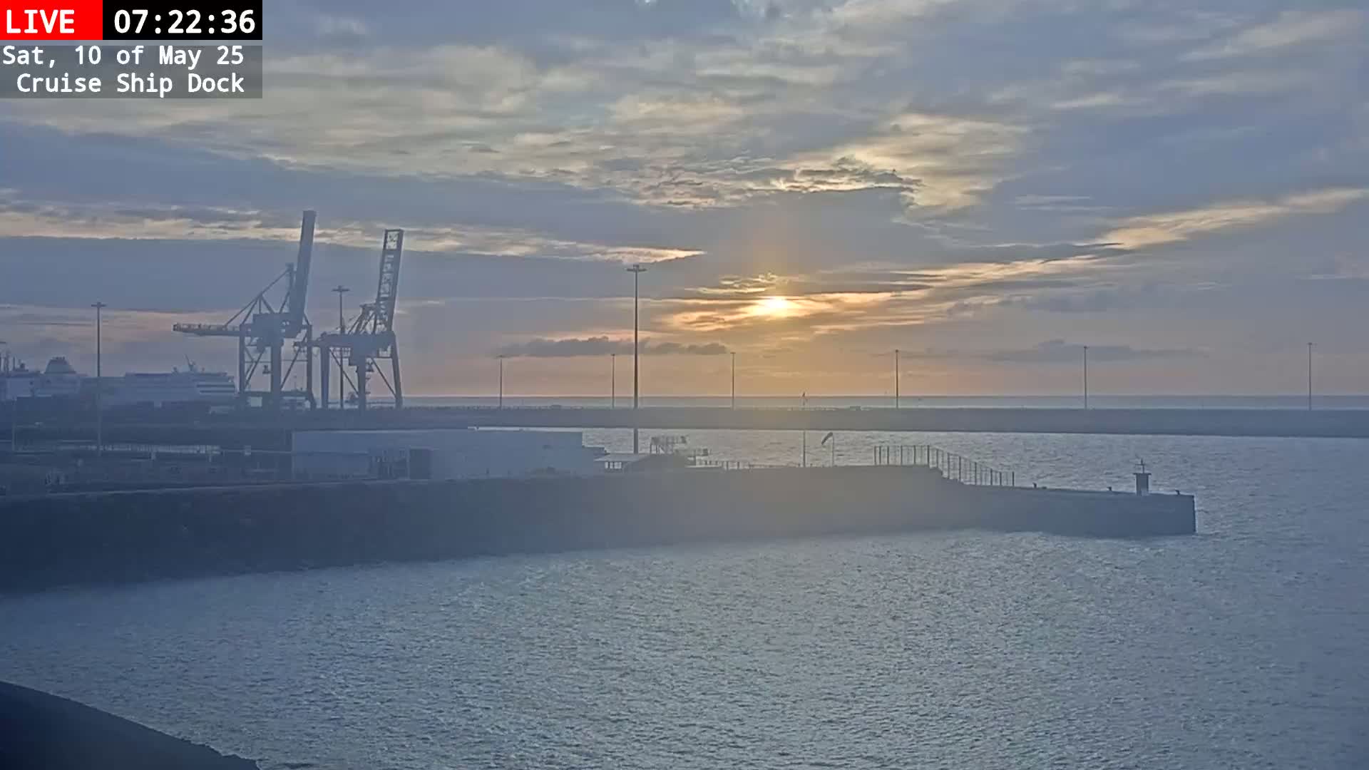 A hazy sunrise casts a warm glow over a calm harbor, where cargo cranes and docked ships are visible under a mostly cloudy sky.