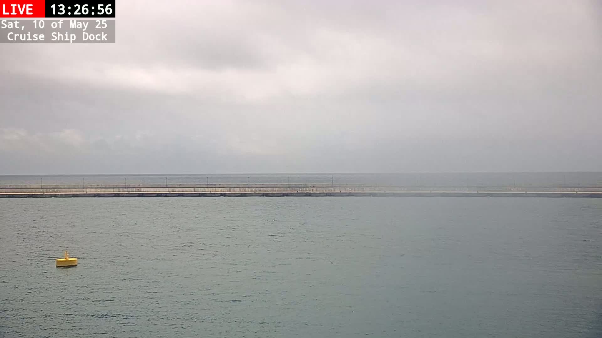 A long, low seawall stretches across a calm, gray body of water under an overcast sky, with a single yellow buoy visible in the foreground.