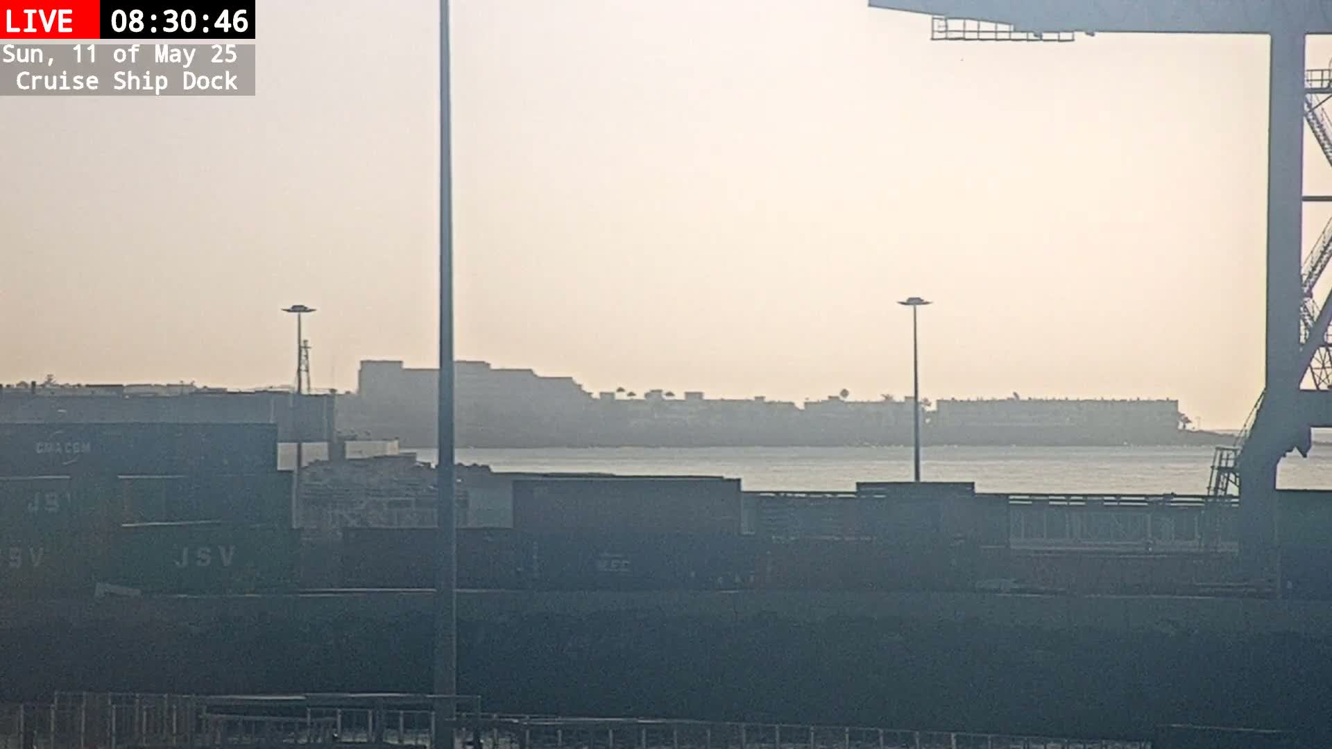 A hazy view of a body of water shows a distant shoreline with buildings and a closer view of a dock with cargo containers and part of a crane.