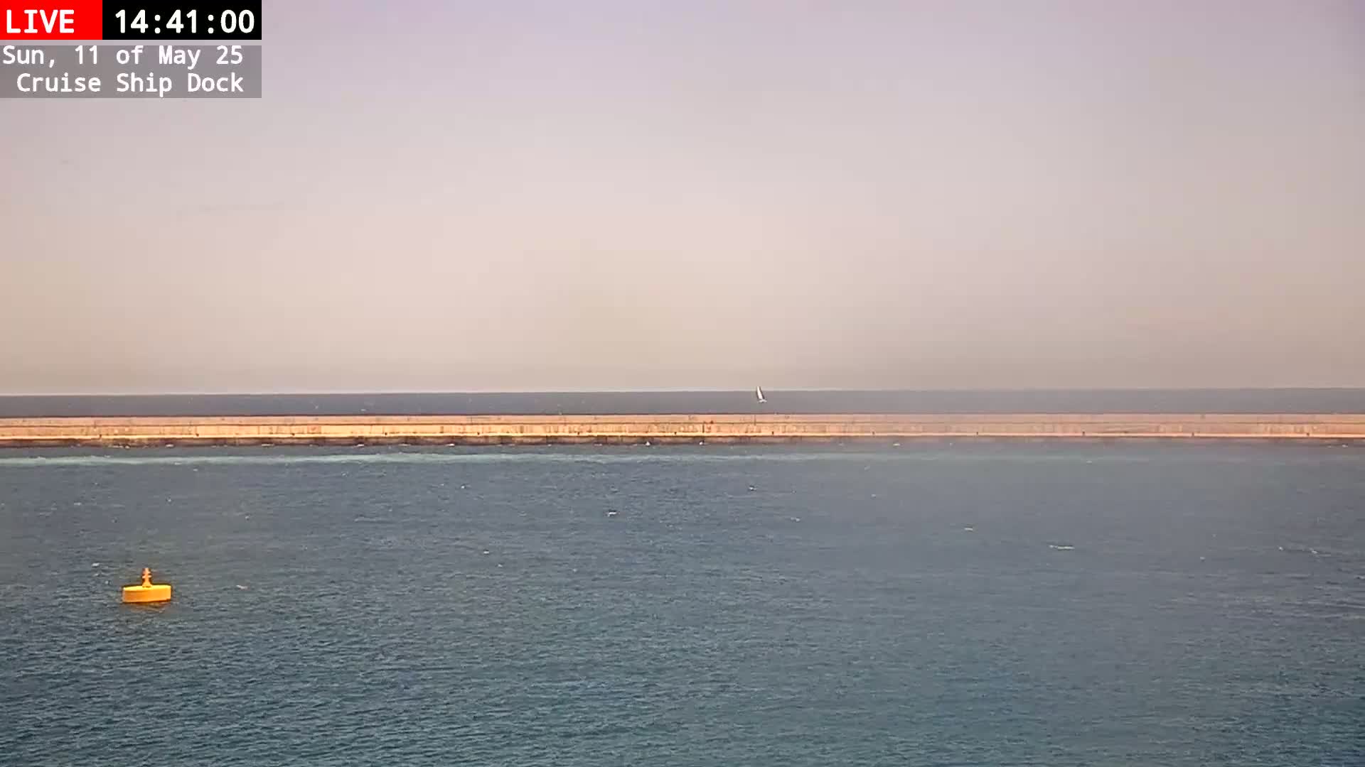 A long, low breakwater stretches across a calm, hazy sea with a single small sailboat visible in the distance, and a yellow buoy near the bottom left.