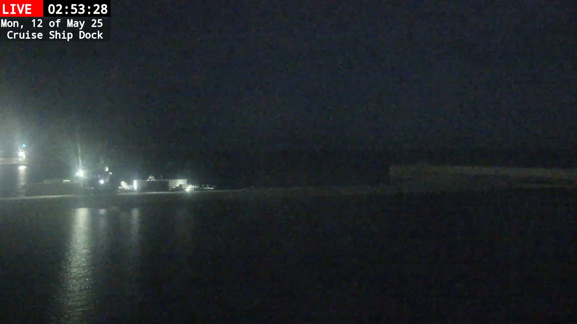 A nighttime view shows a dark body of water reflecting dim lights from a dock and buildings on the far shore.