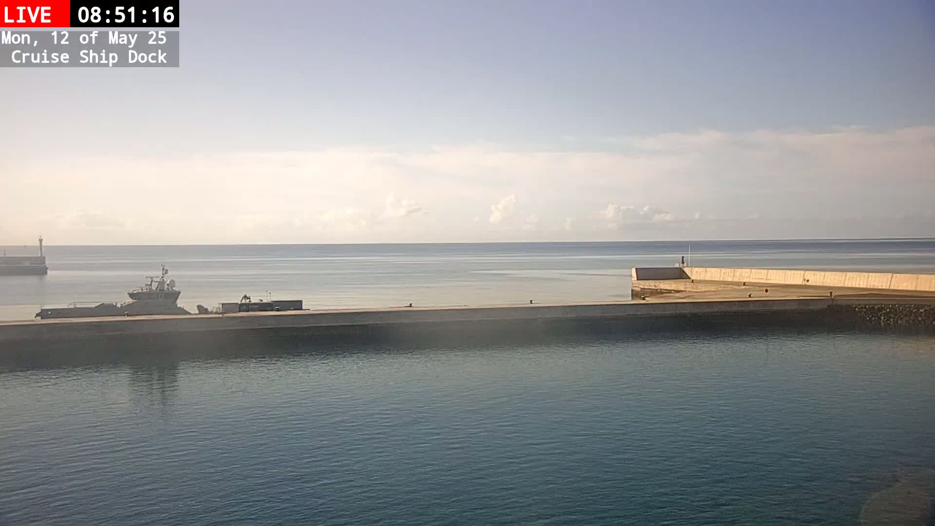 A calm, partly cloudy day reveals a harbor with a docked ship and a long concrete pier extending into the ocean.