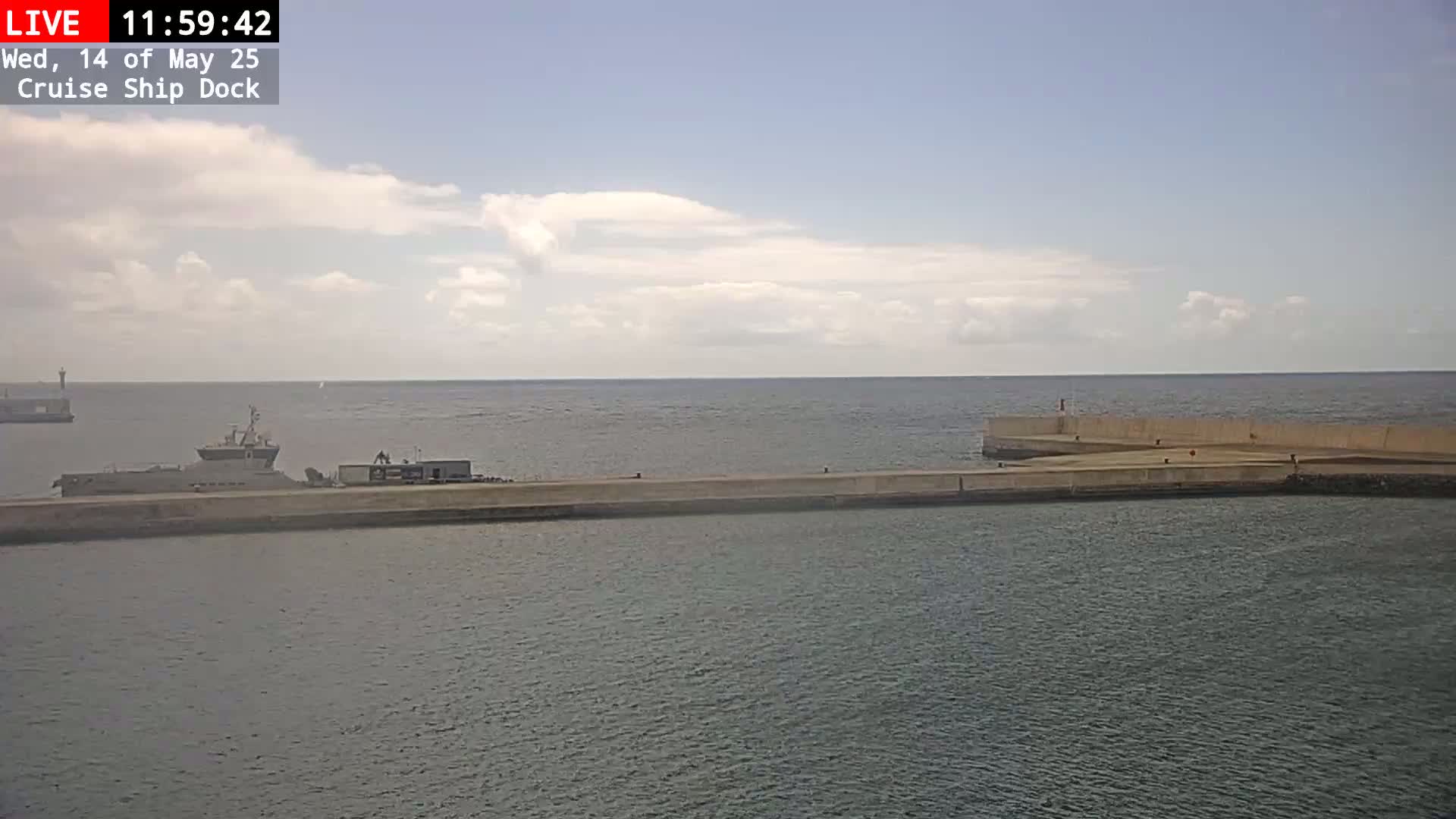 A gray ship is docked at a concrete pier on a partly cloudy day, with calm water surrounding it.