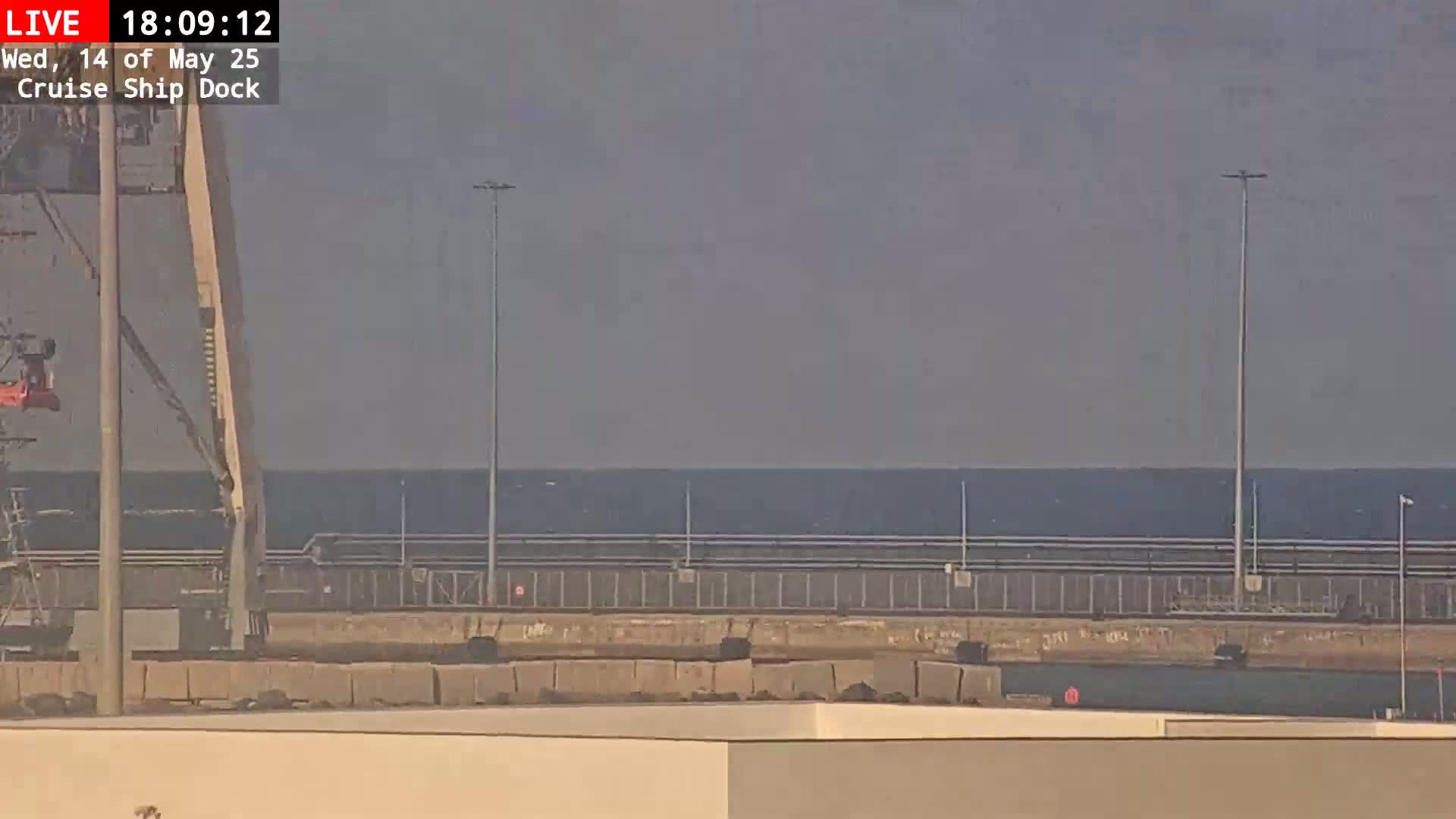 A mostly clear sky overlooks a calm body of water behind a security fence and industrial structures at a cruise ship dock.