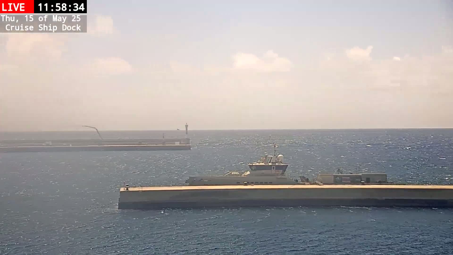 A long, low-lying vessel is moored at a dock in hazy, sunny conditions.