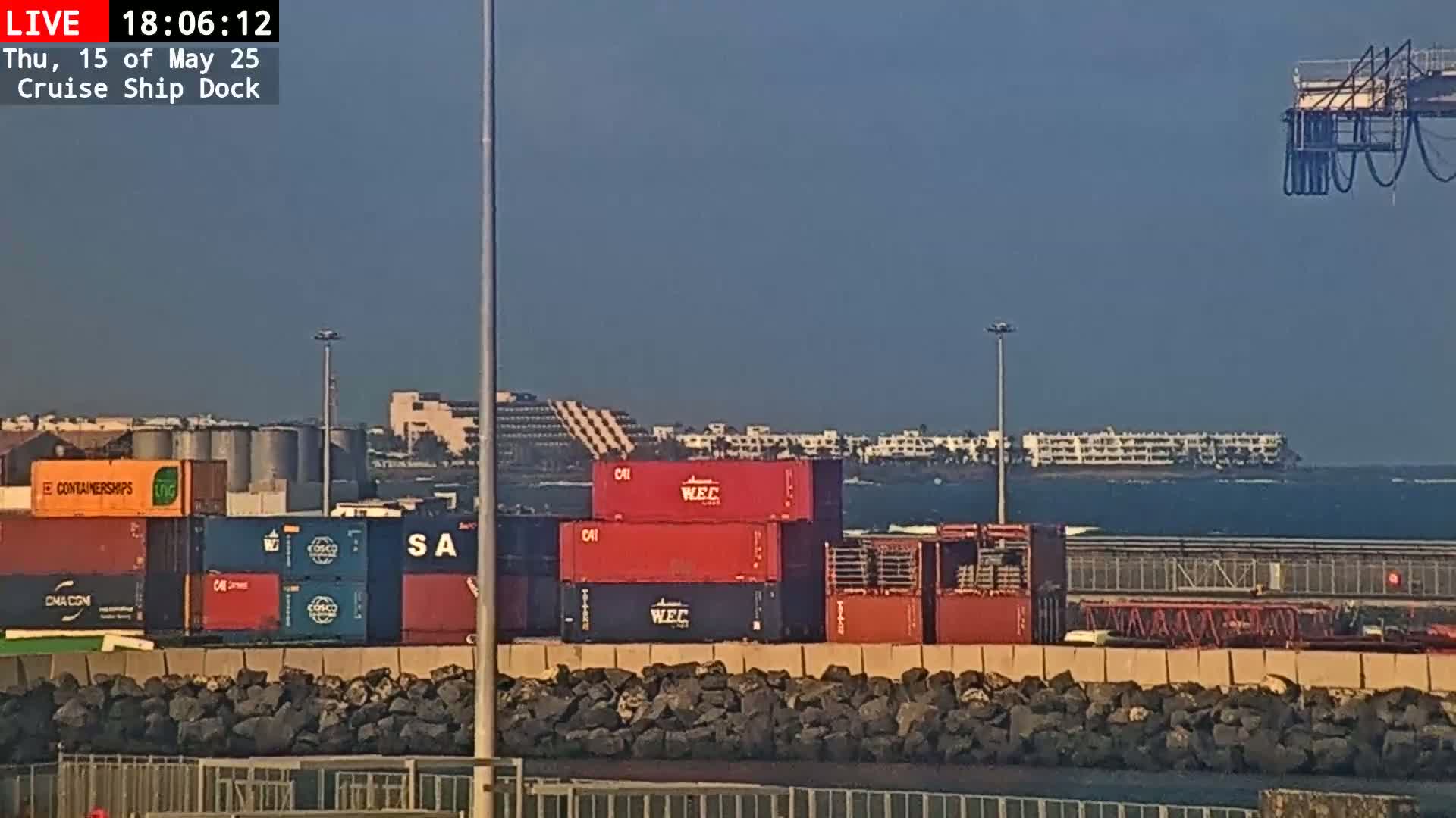 A sunny day at a cruise ship dock shows numerous shipping containers stacked near a rocky wall, with a distant coastline and buildings visible across calm water.