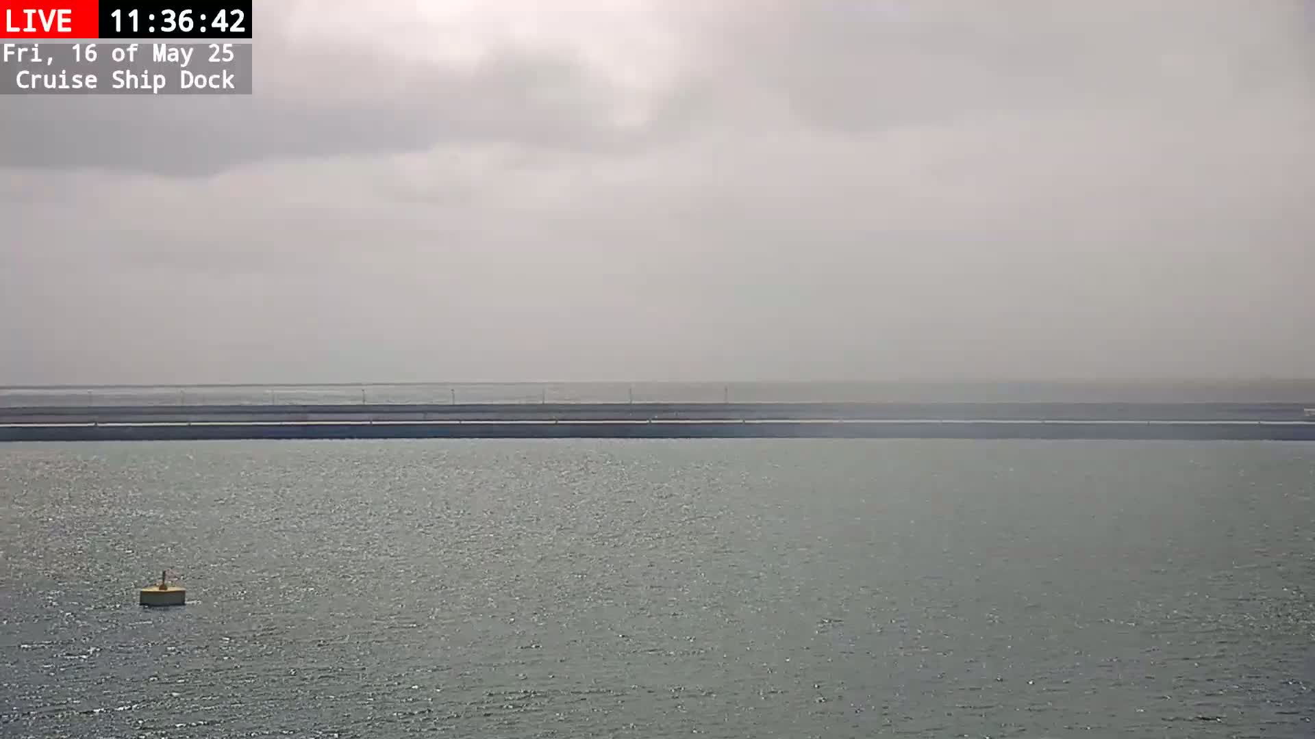 A body of water stretches out under a hazy, overcast sky, with a single buoy visible in the foreground and a long, dark structure in the distance.