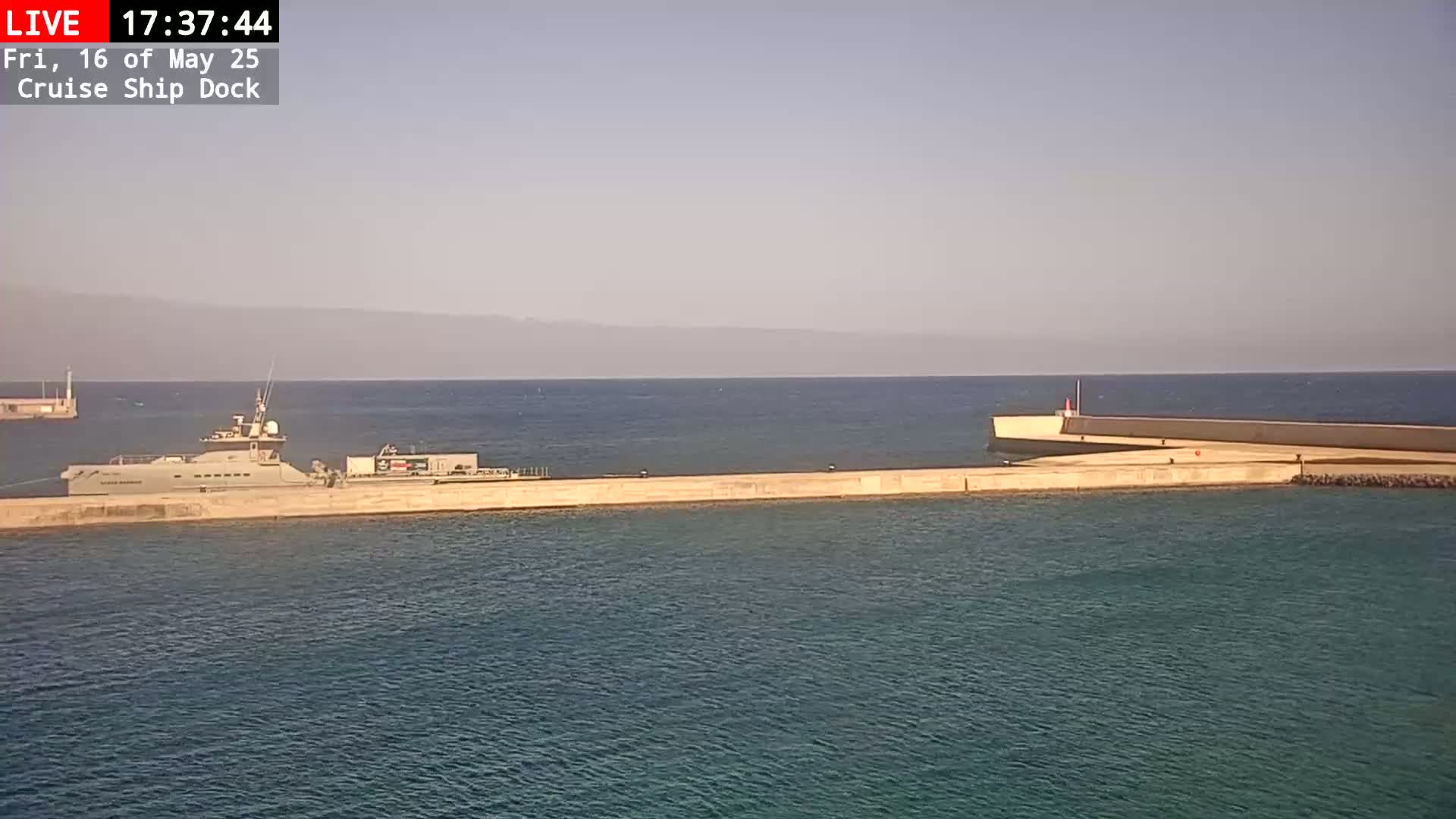 A long, light-colored vessel is docked at a concrete pier on a calm, hazy day, with mountains visible in the distance.