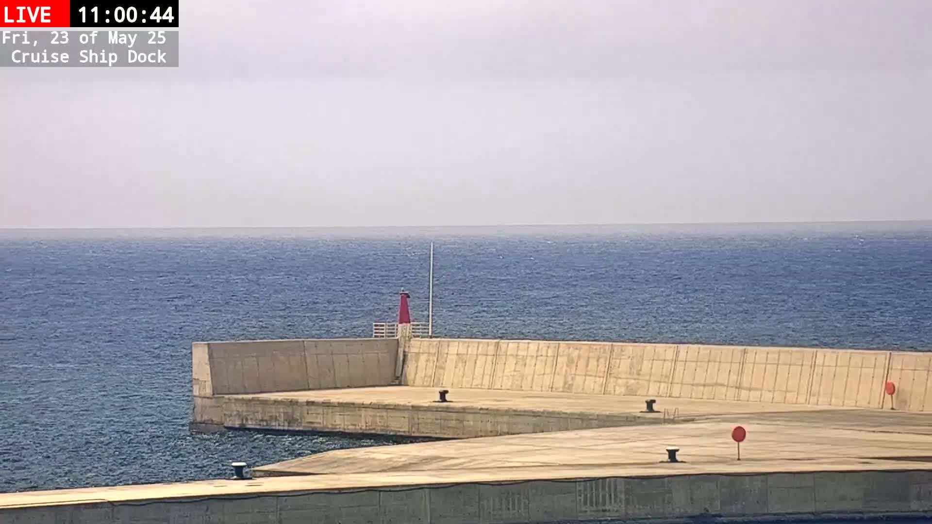 A concrete dock extends into a calm, blue ocean under a hazy sky, featuring a small red lighthouse.