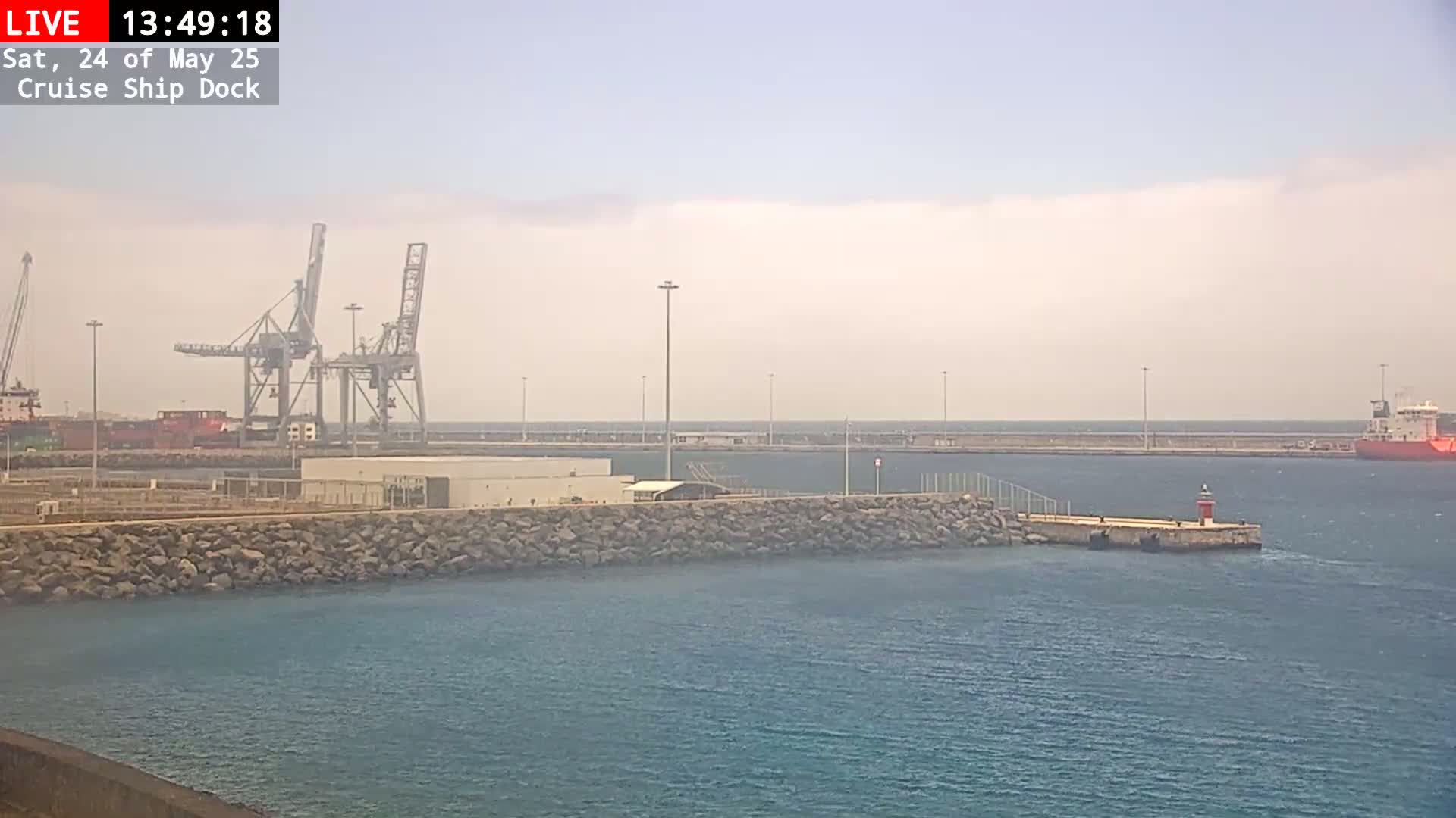 A hazy day shows a harbor with cargo cranes, a breakwater, and a ship in the distance.