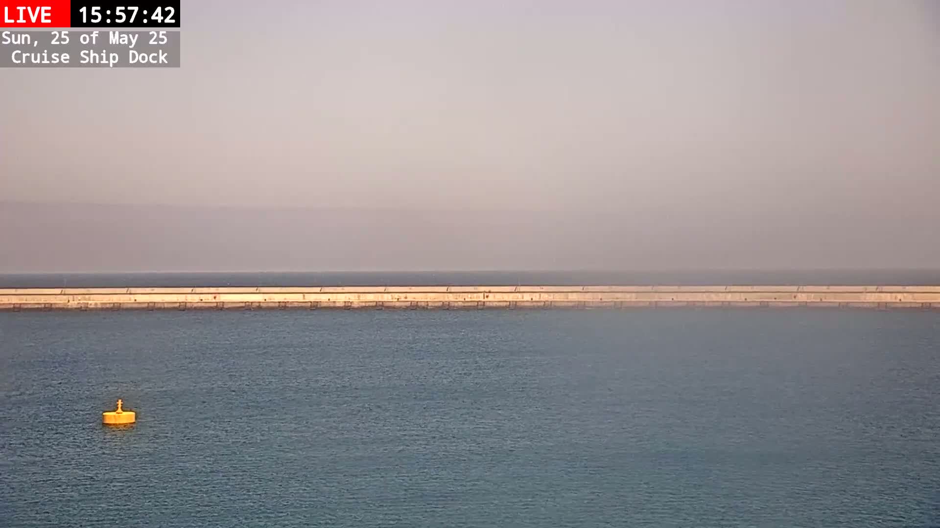 A long, low seawall stretches across a calm, hazy body of water, with a single yellow buoy visible in the foreground.