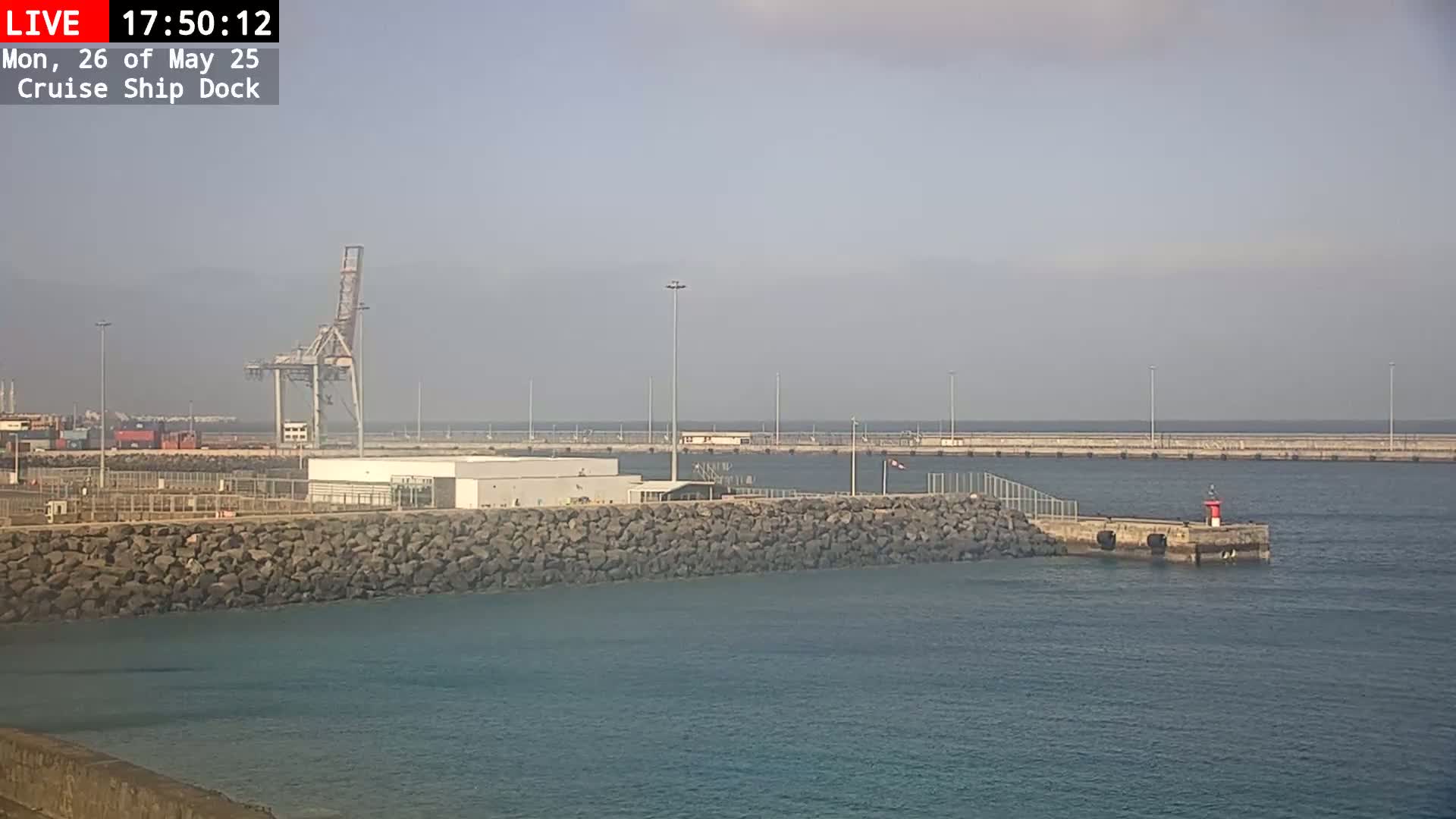 A hazy, sunny day at a cruise ship dock shows a large crane and a rock seawall separating calm water from a port area.