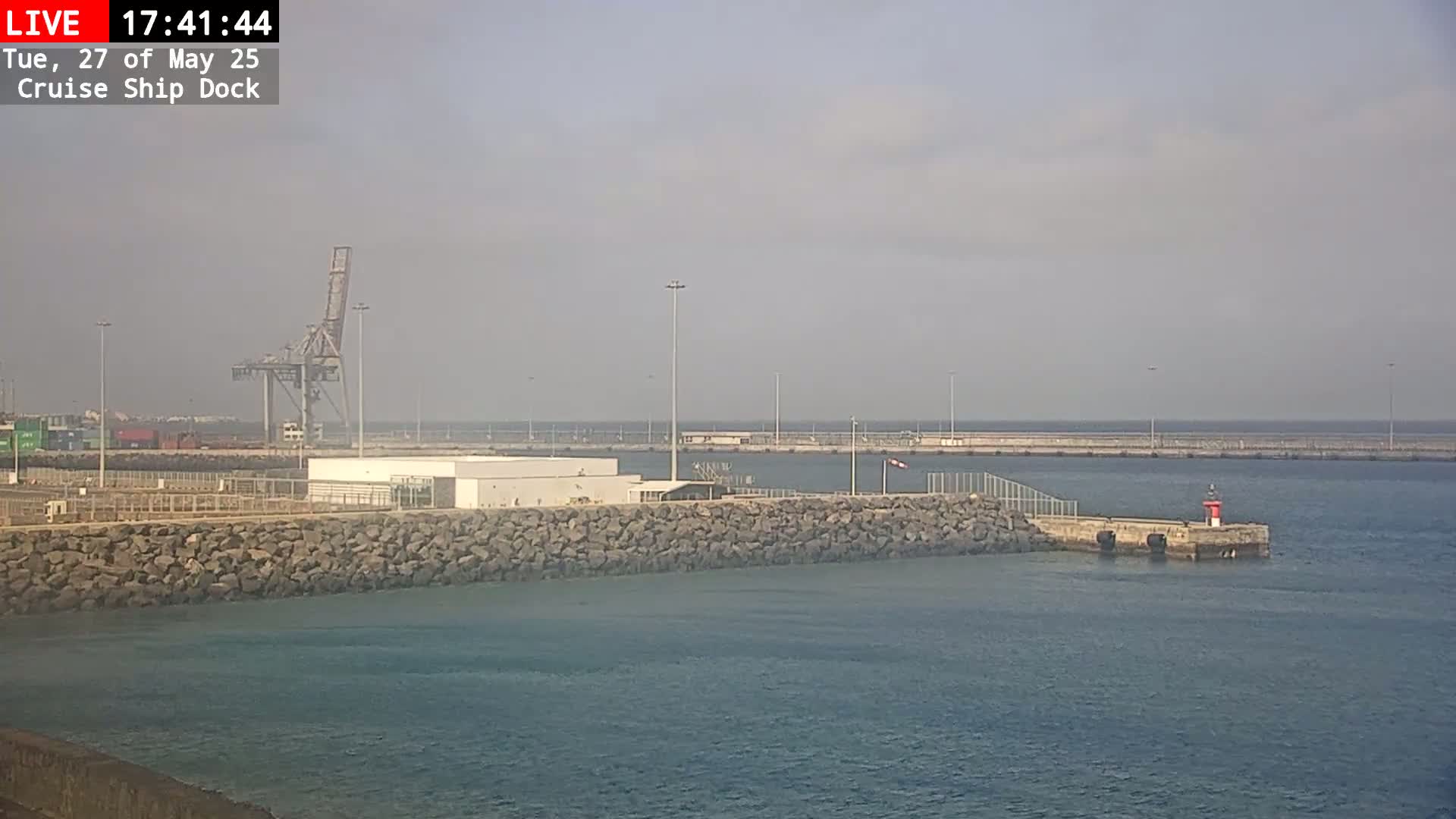 A hazy view of a cruise ship dock shows a portion of a harbor with a crane, buildings, and a small lighthouse on a pier.