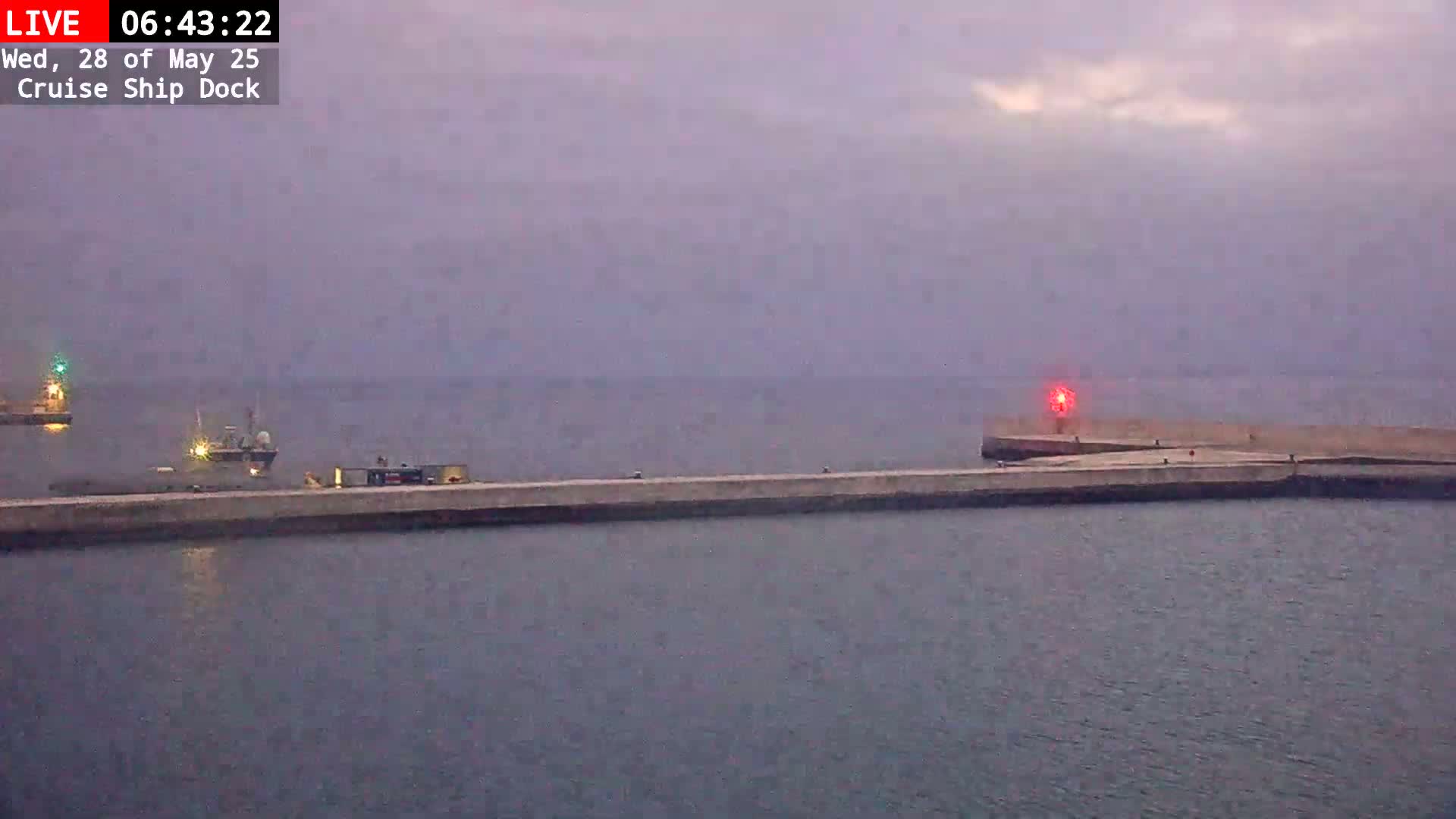 A long concrete pier extends into a calm, overcast body of water, with two small ships visible in the distance.