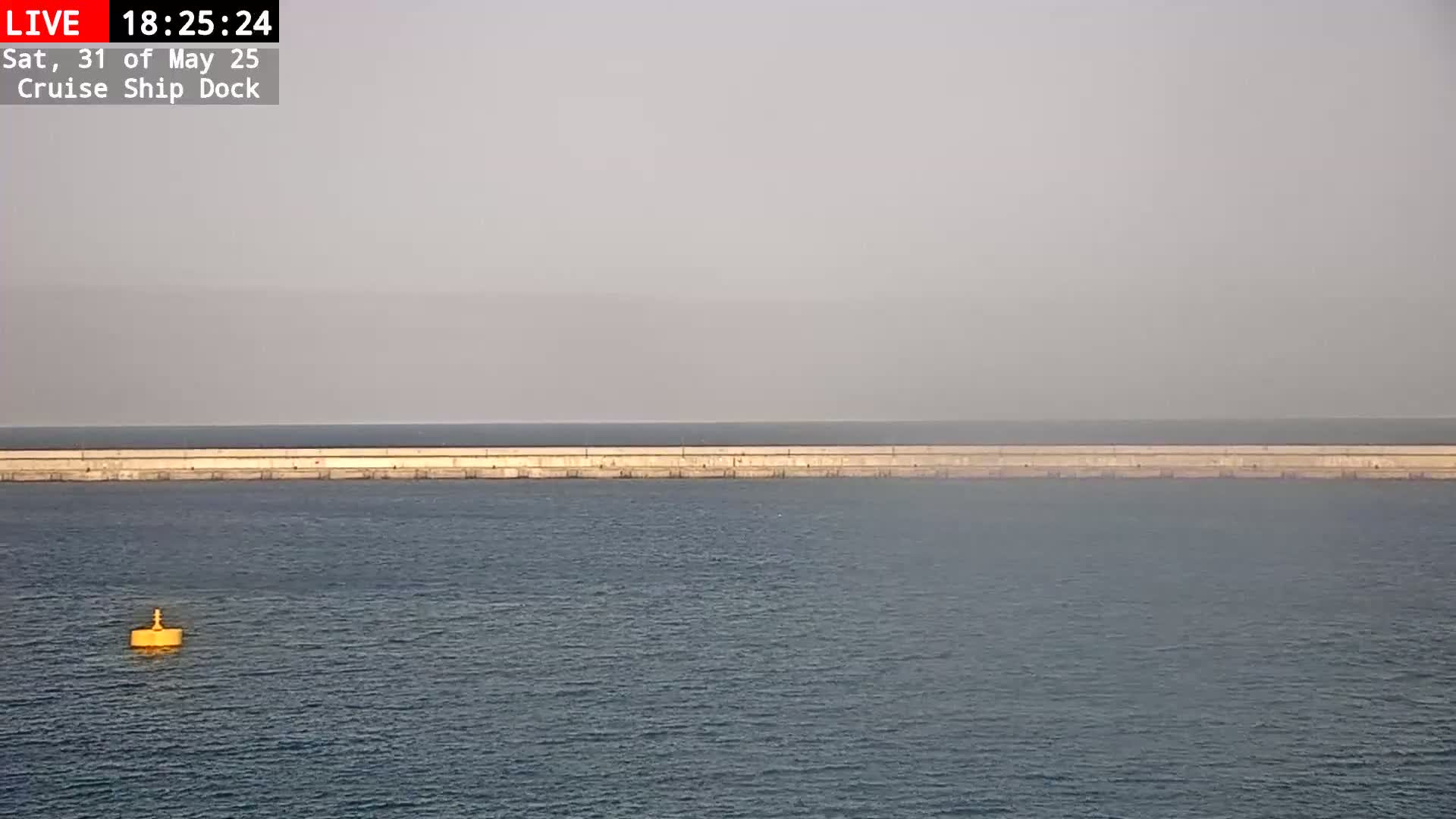 A long, low seawall stretches across a hazy, calm body of water, with a single yellow buoy visible in the foreground.