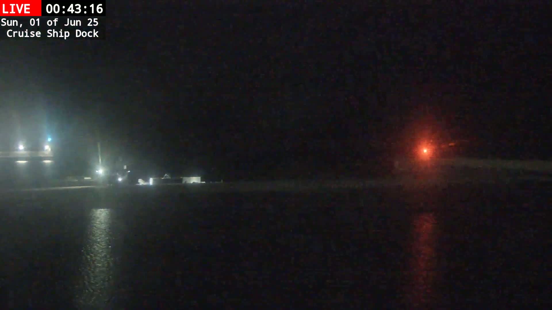 A nighttime view of a dark body of water reflecting a faint red light, with dimly lit structures visible on the shore in the distance.