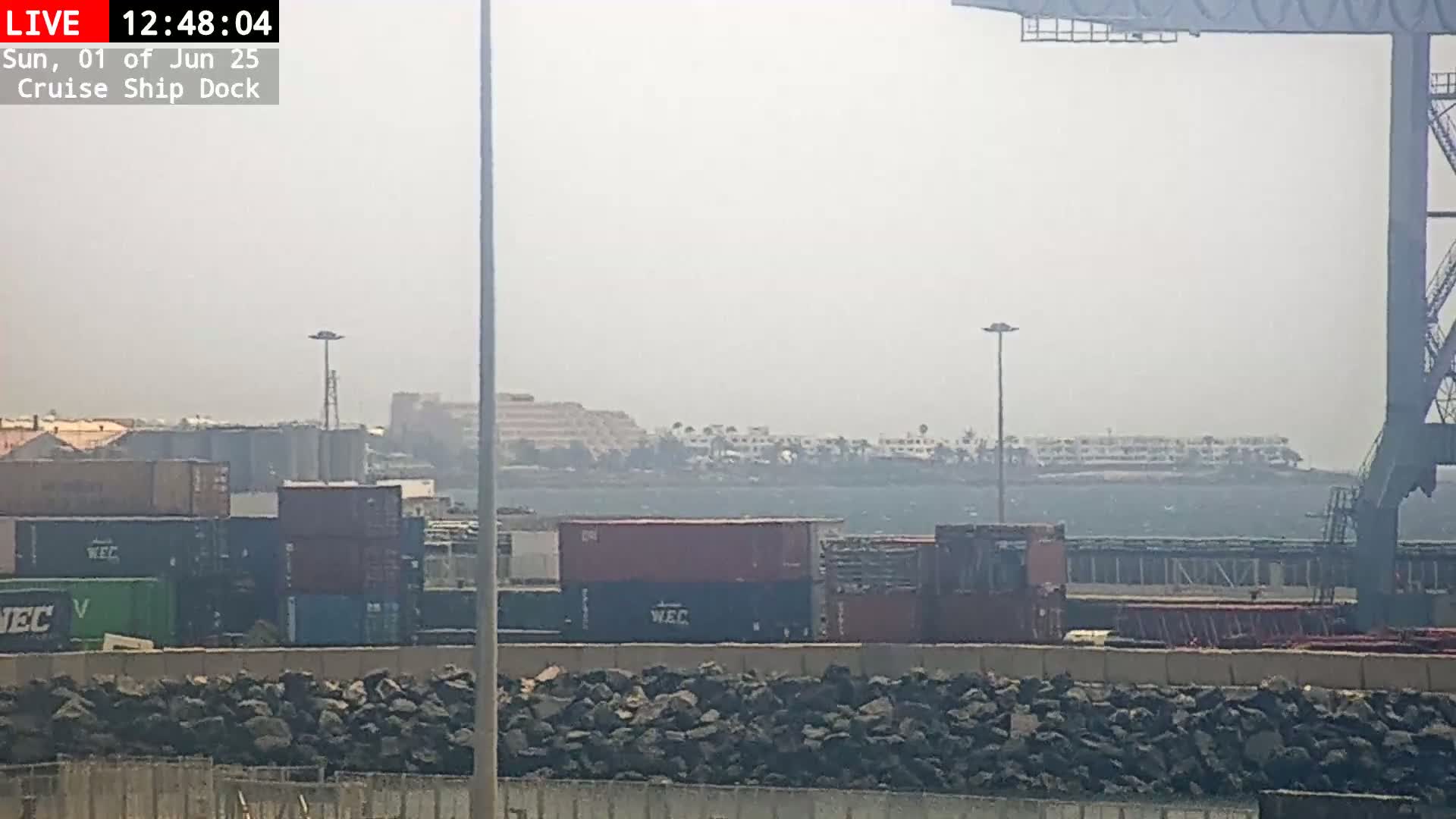 A hazy view of a cruise ship dock shows shipping containers, a rocky shoreline, and a distant line of buildings.