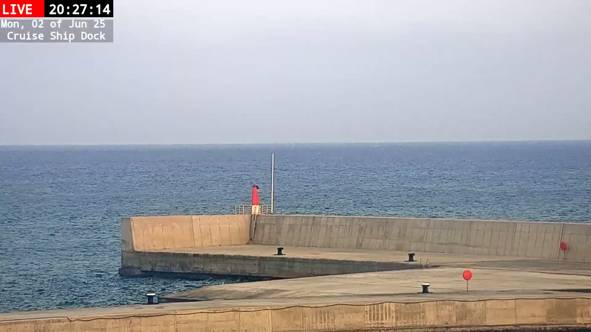 A concrete pier extends into a calm, blue sea under a hazy sky, with a small red lighthouse visible on the pier.