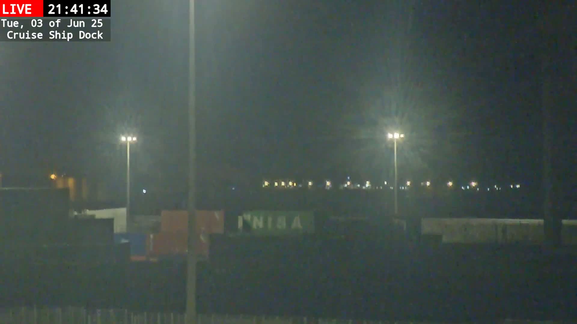At night, under dark, hazy conditions, a dimly lit dock area shows several containers and tall light poles.