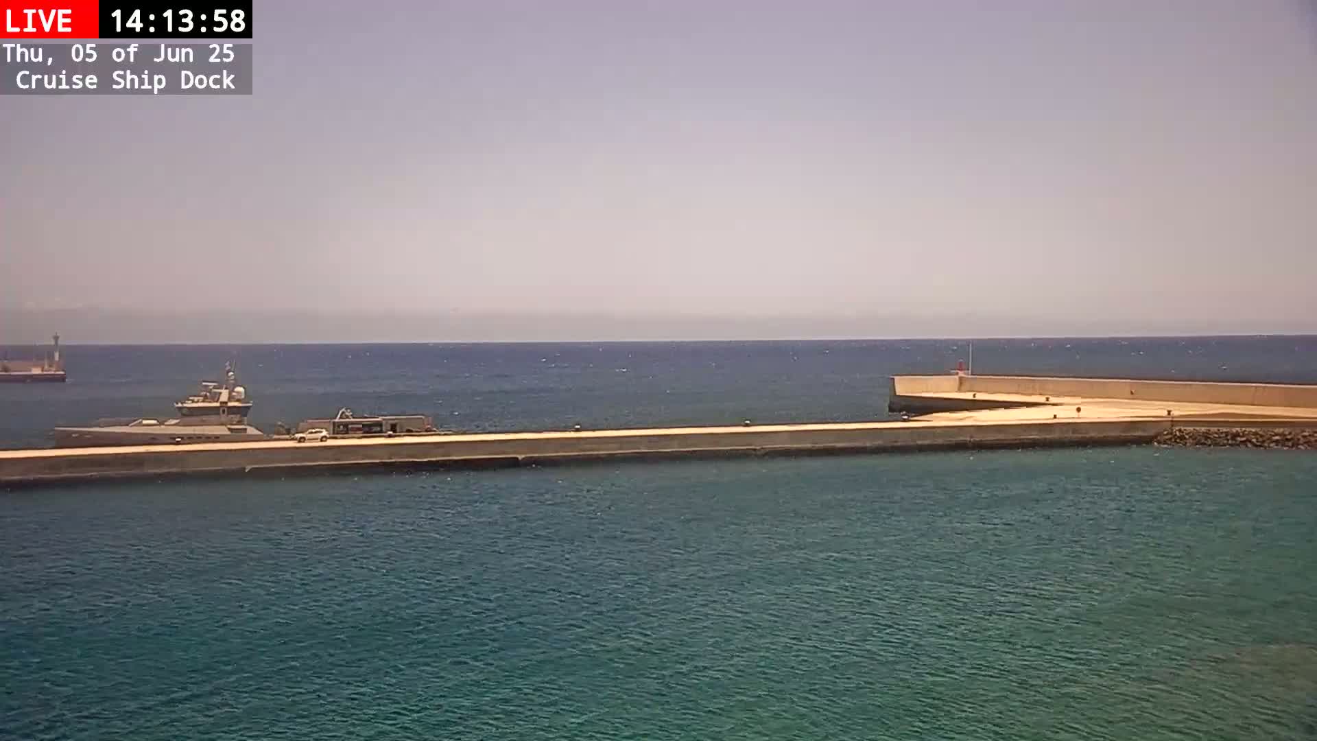 A large vessel is docked at a long concrete pier extending into a calm, teal sea under a hazy sky.