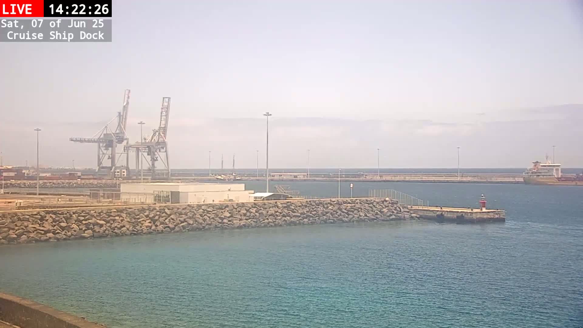 A hazy harbor scene shows a cruise ship dock with several cranes, a stone wall, and a large ship in the distance.