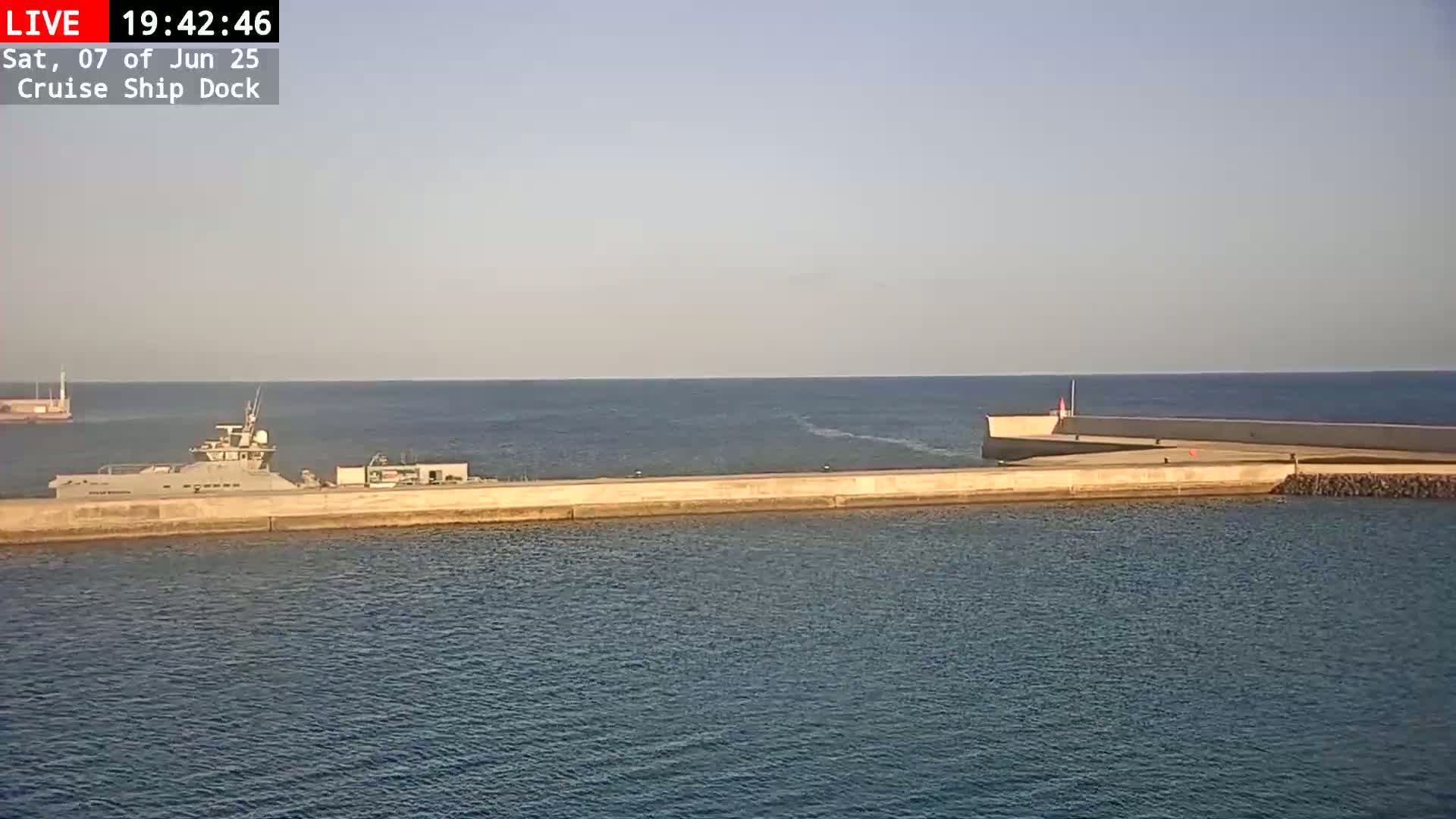 A large gray ship is docked at a concrete pier on a calm, sunny day.