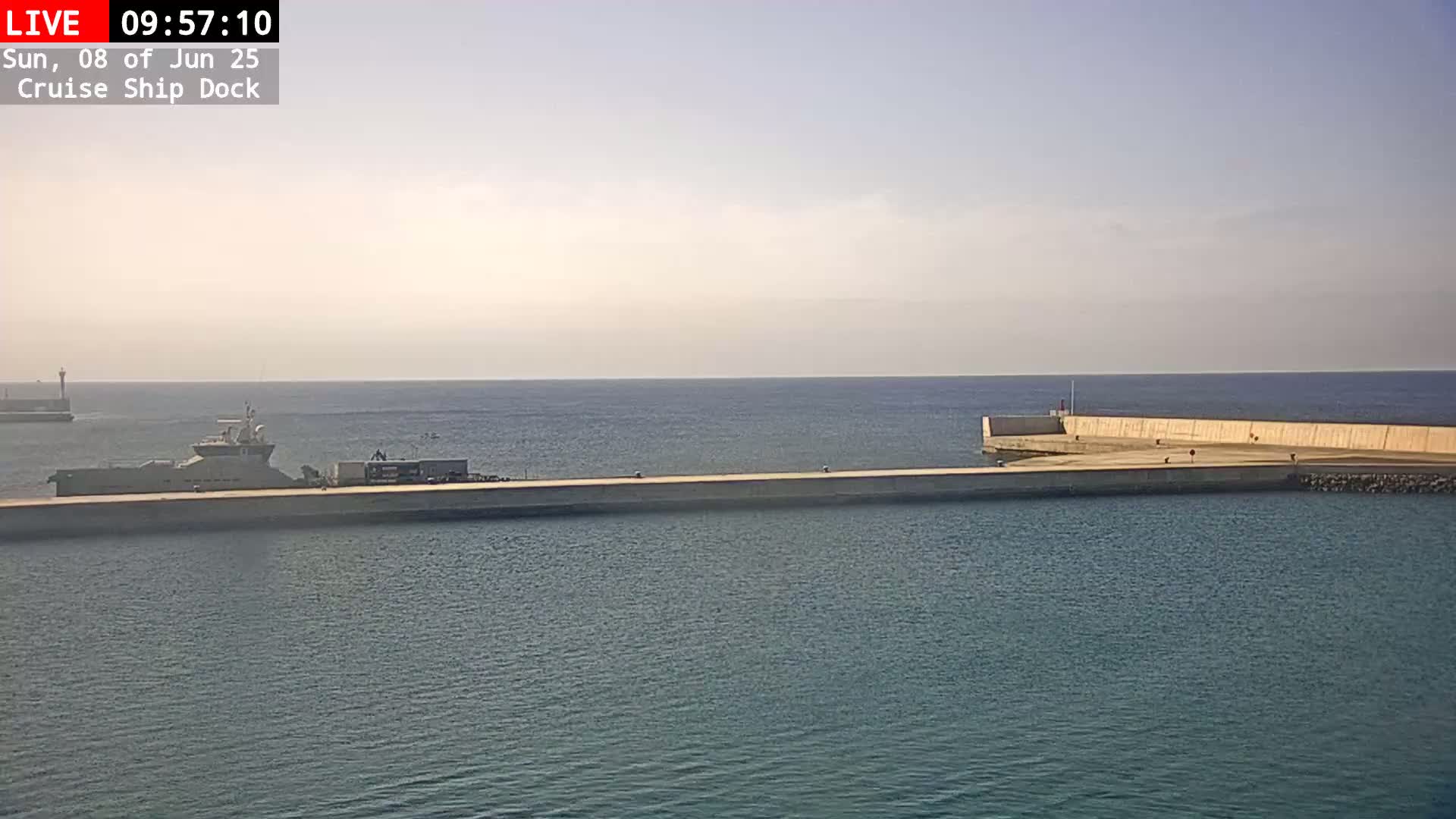 A long, light-colored dock extends into a calm, blue ocean on a partly sunny day, with a ship docked near the end of it and another in the distance.