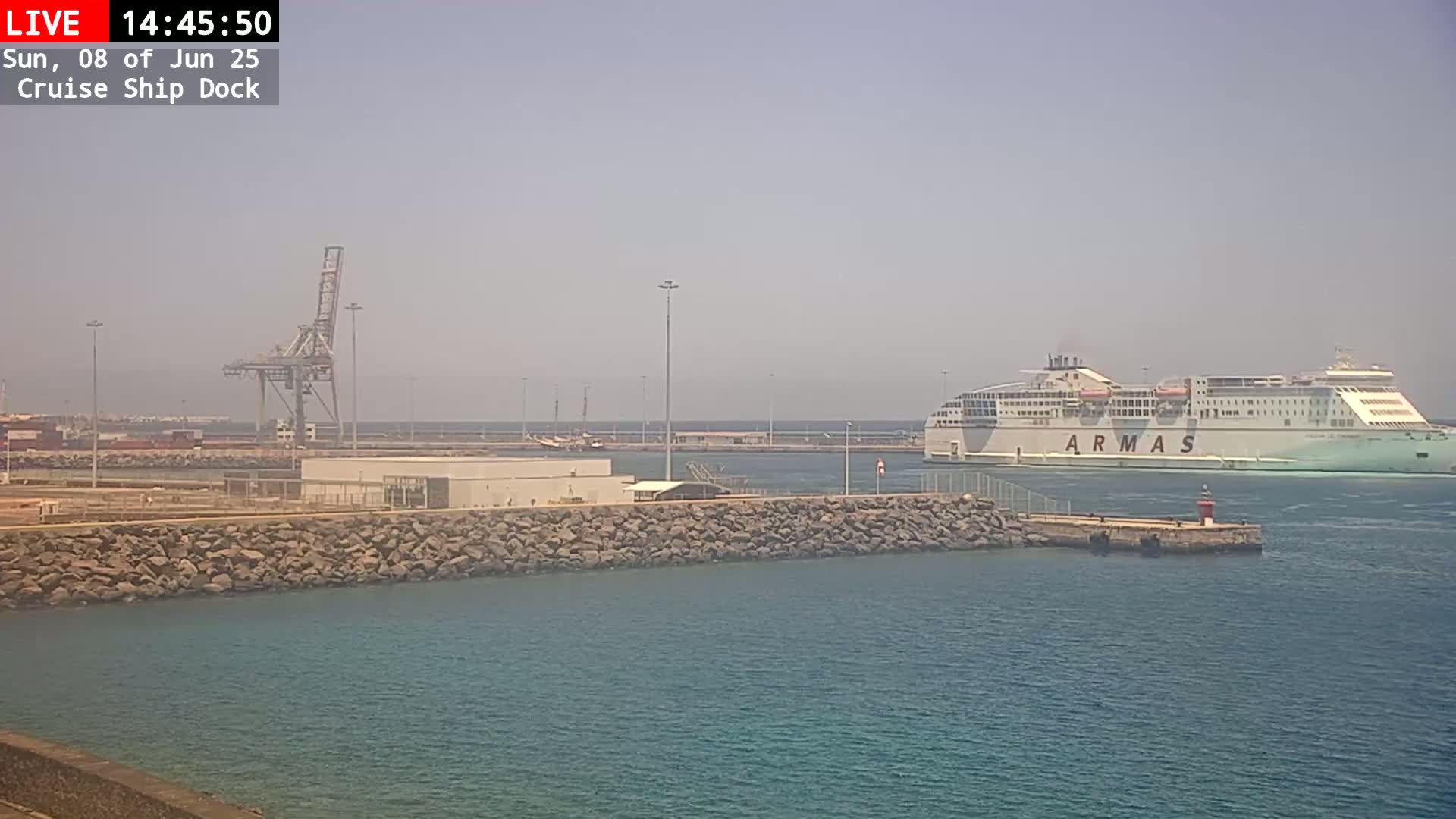 A large ferry approaches a harbor on a hazy day, passing a smaller pier and a large crane.