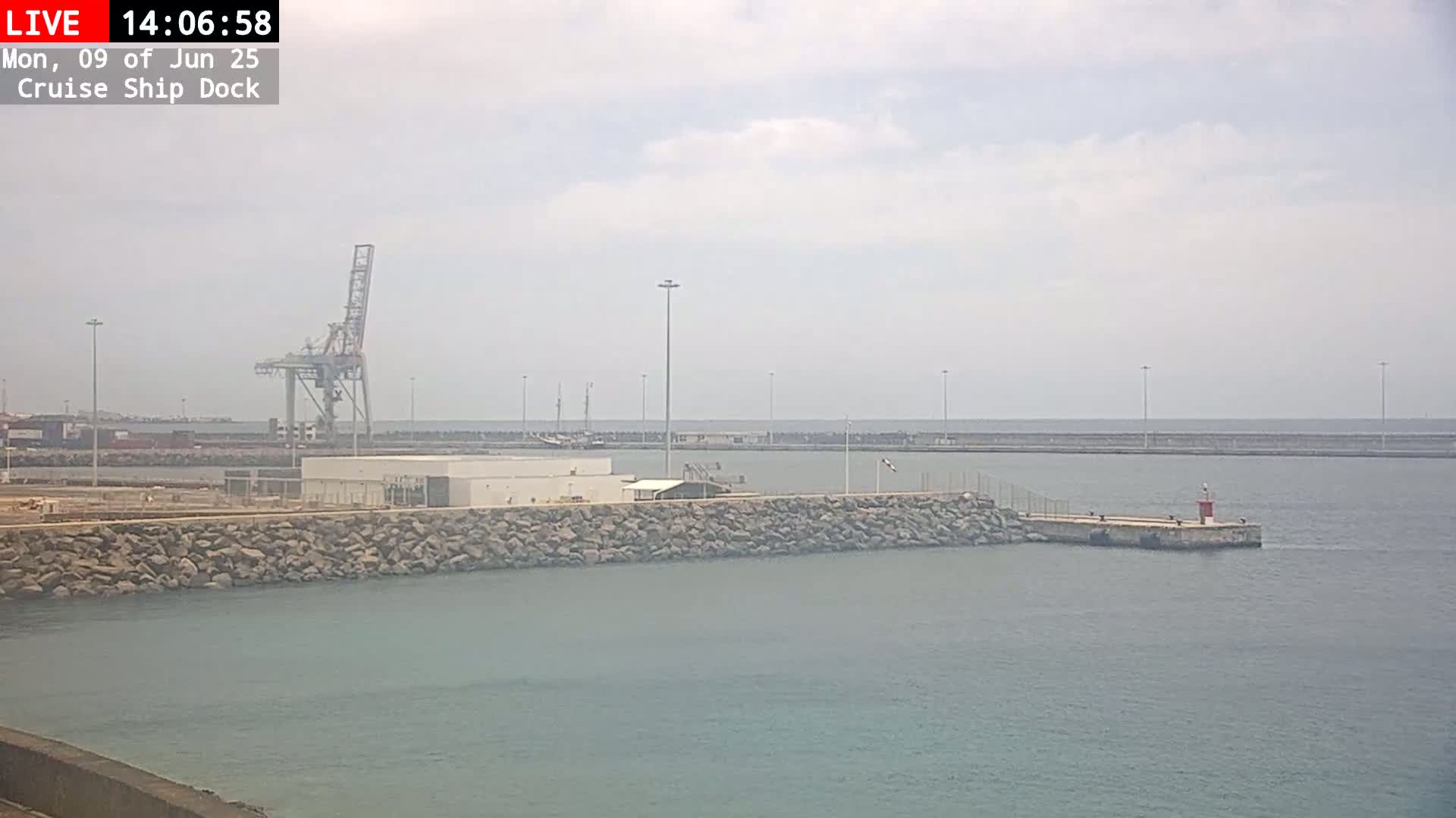 A partly cloudy day shows a cruise ship dock with a large crane, a rock wall, and calm water.