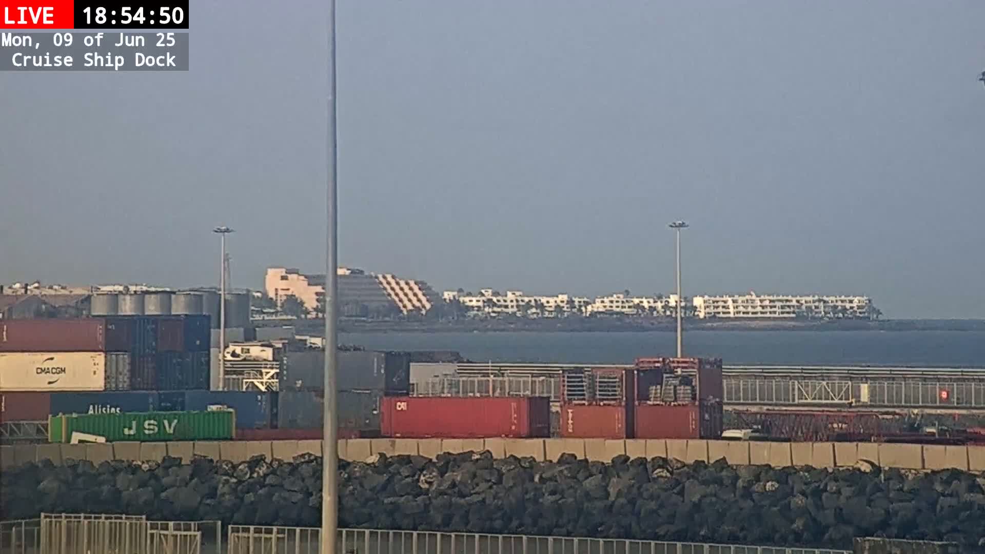 A mostly clear day reveals a cruise ship dock with shipping containers, a rock barrier, and a distant shoreline featuring buildings.