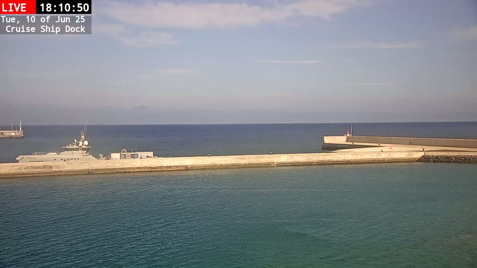 A gray ship is docked at a concrete pier extending into a calm, blue ocean under a mostly clear sky.