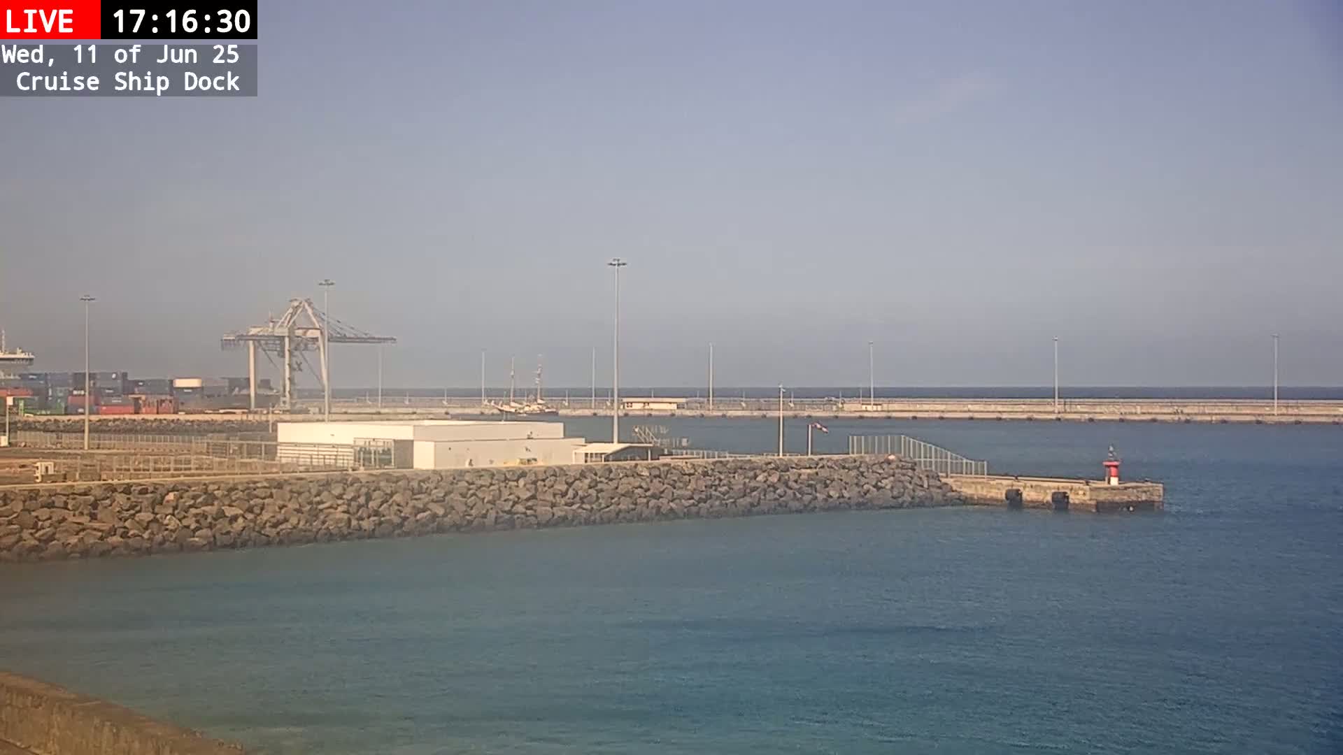 A hazy, sunny day shows a cruise ship dock with cargo containers, cranes, and a small lighthouse on a jetty extending into calm blue water.