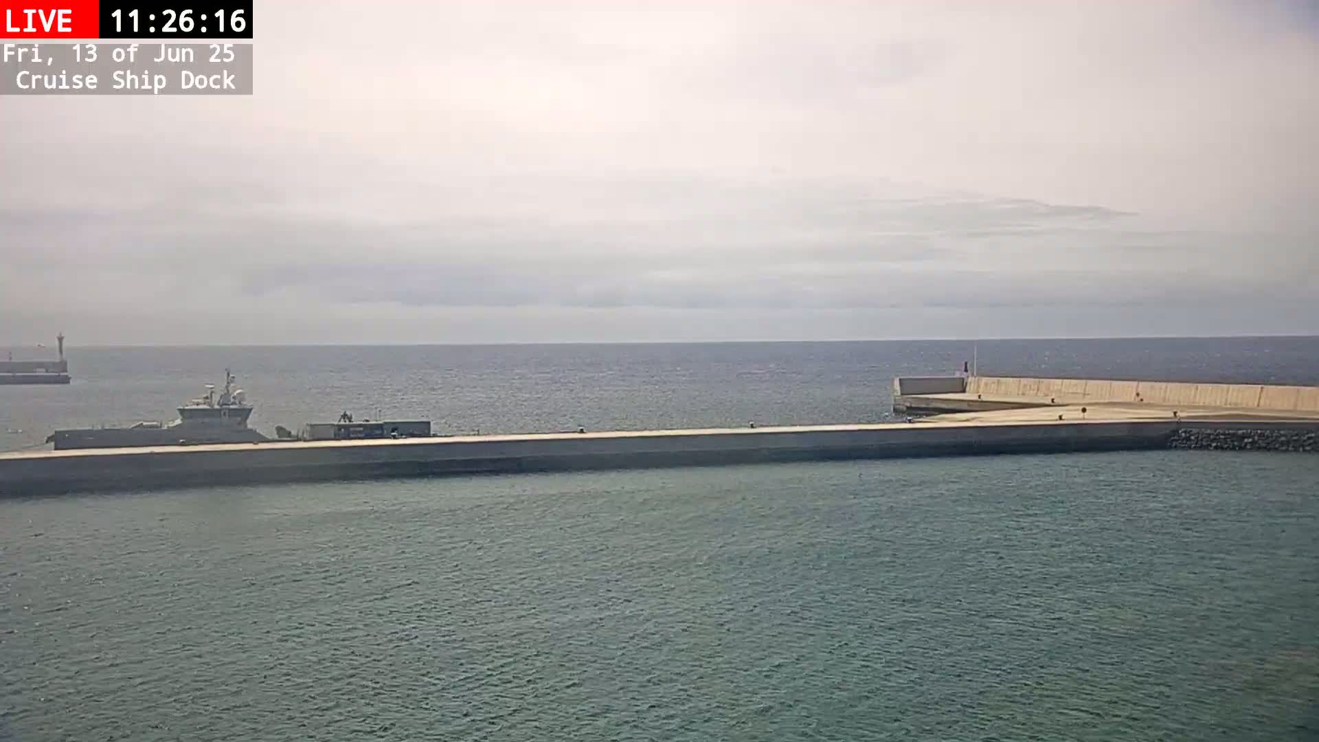 A gray ship is docked at a long concrete pier extending into a calm, overcast ocean.
