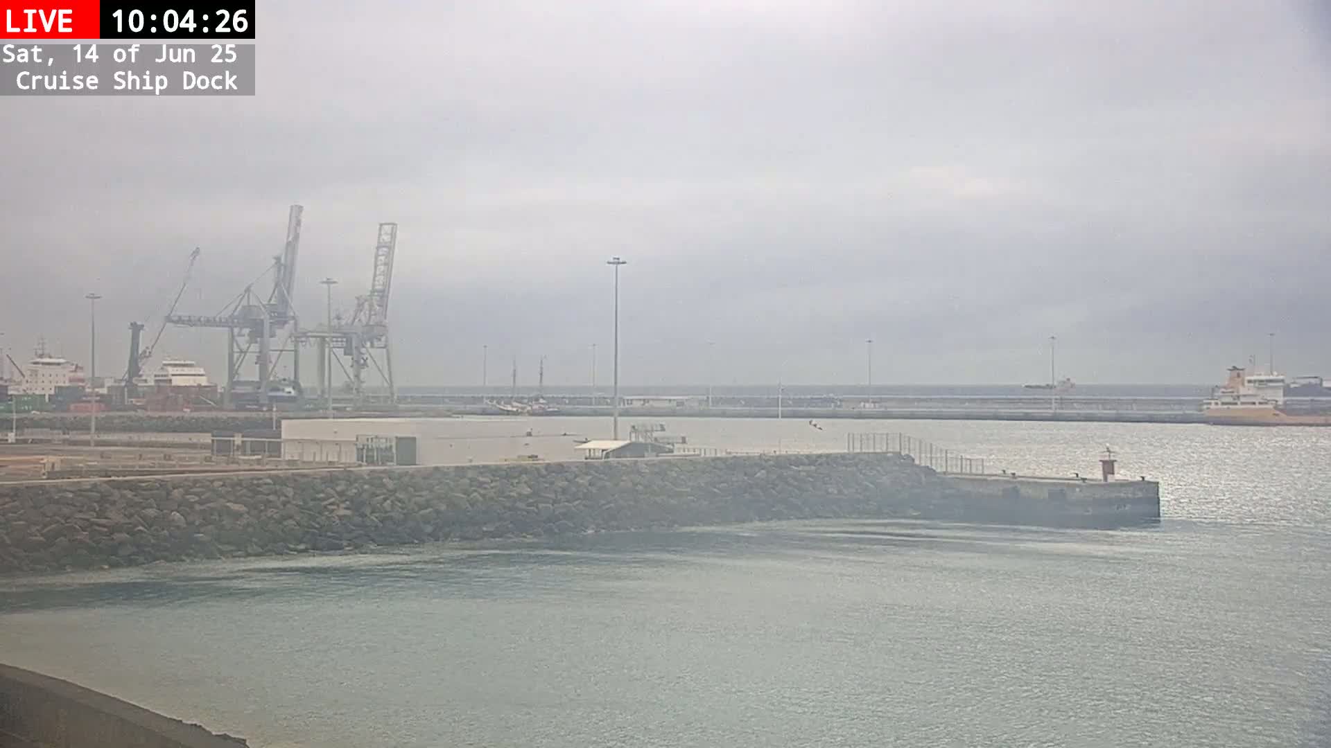 A cruise ship dock is shown on an overcast day, with several cargo ships and cranes visible in the harbor.