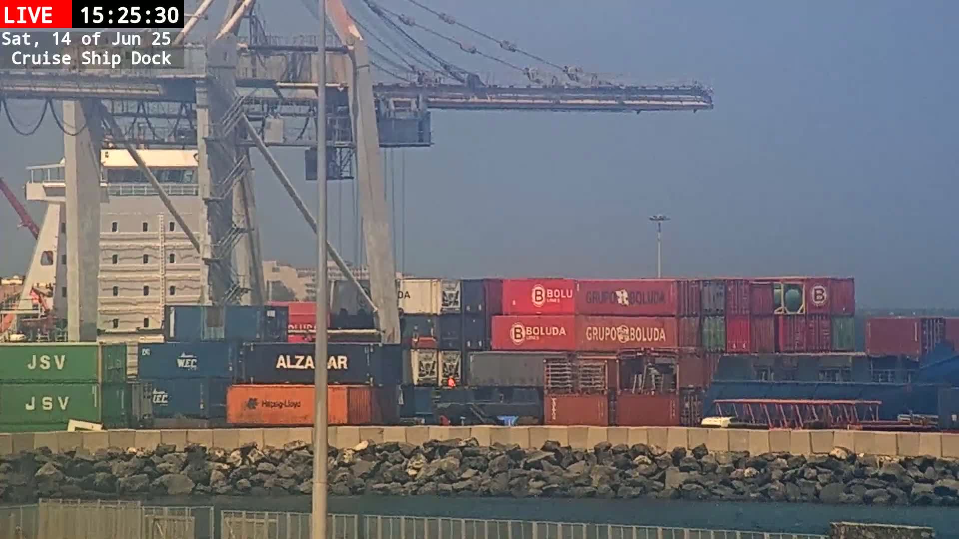 A cruise ship is docked at a port under hazy, sunny conditions, with numerous shipping containers visible on the dock and the ship.