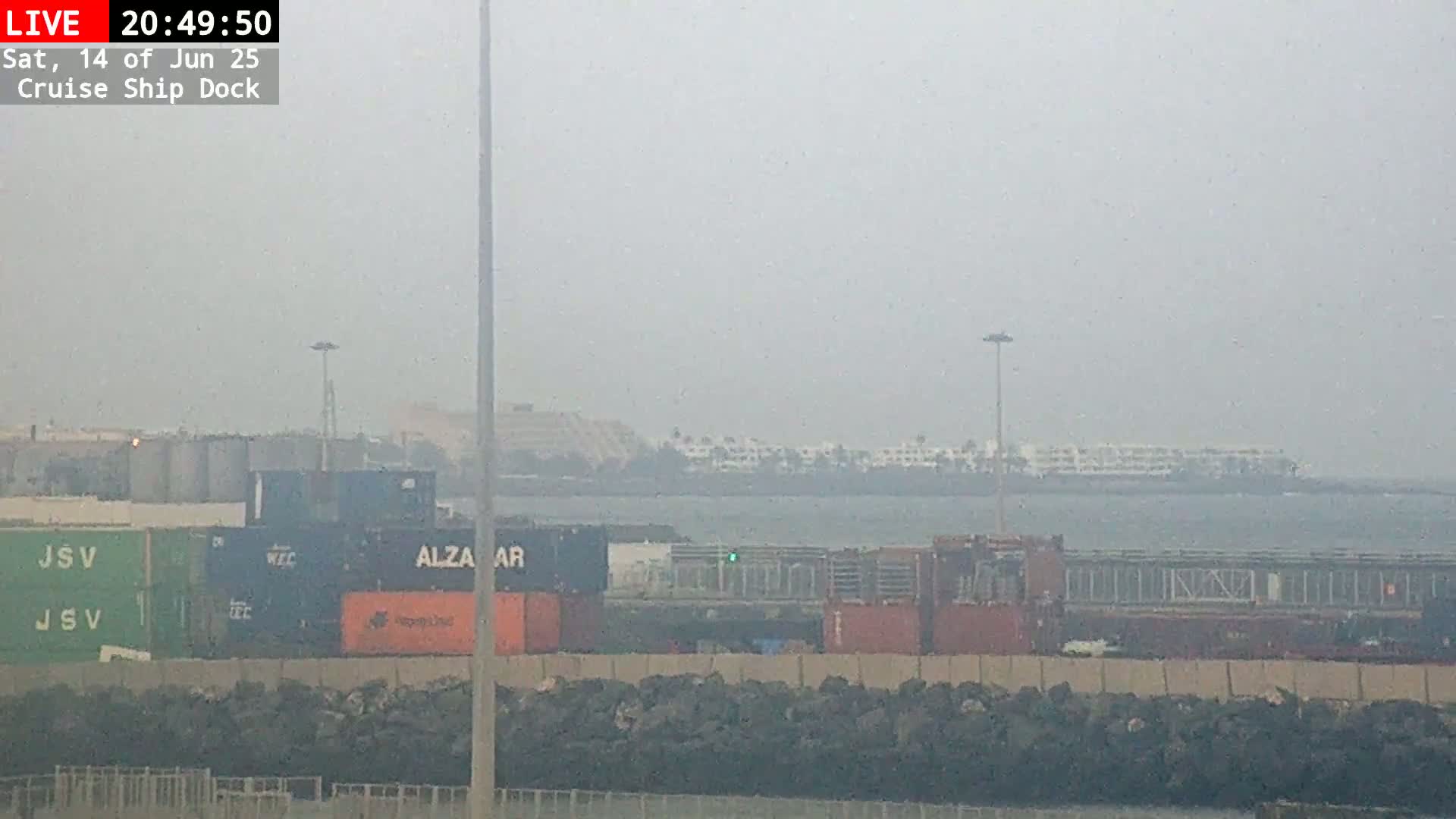 A cruise ship is docked at a port under hazy, sunny conditions, with numerous shipping containers visible on the dock and the ship.
