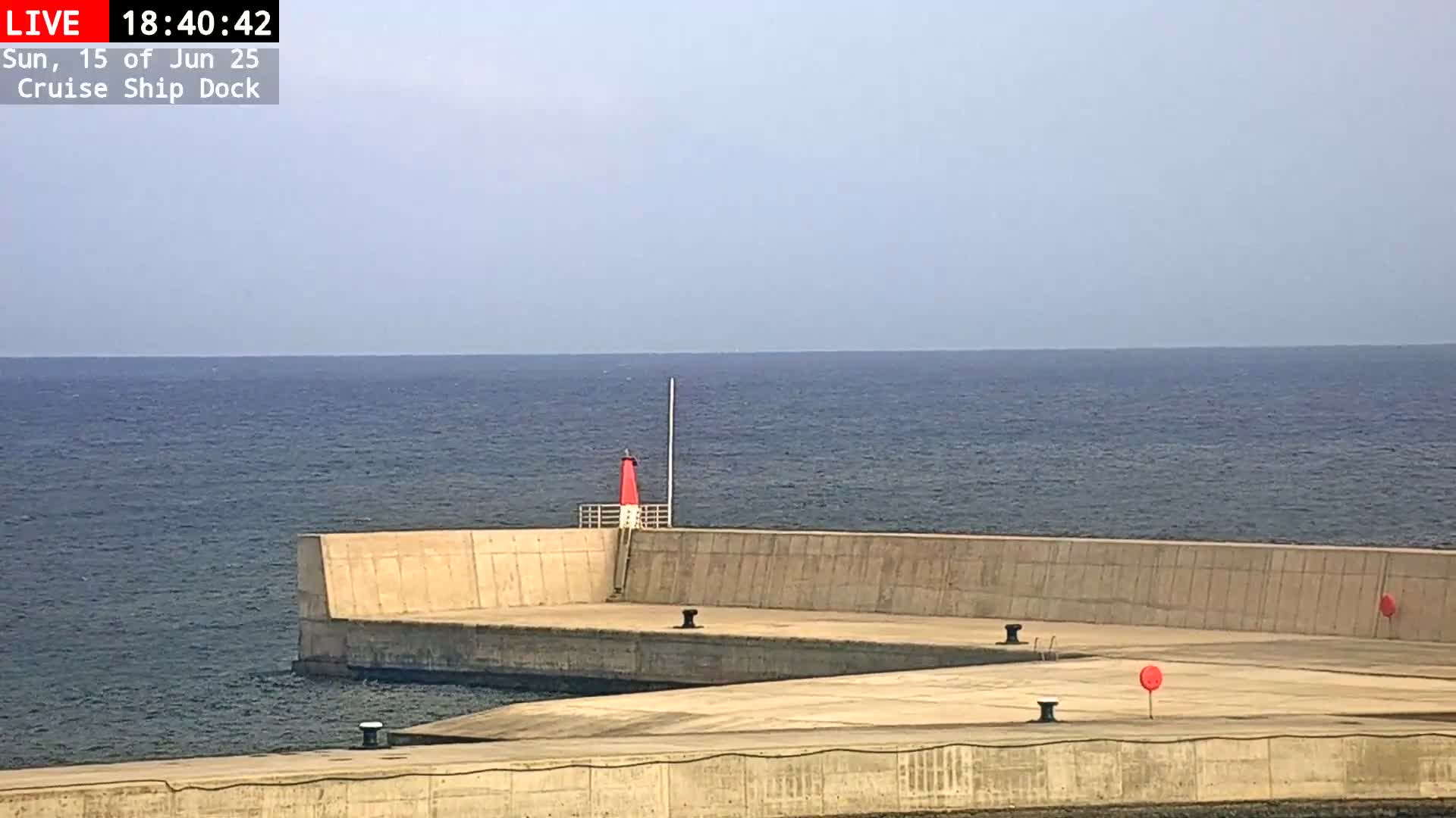 A concrete dock extends into a calm, blue ocean under a clear sky, featuring a small red lighthouse and several red buoys.