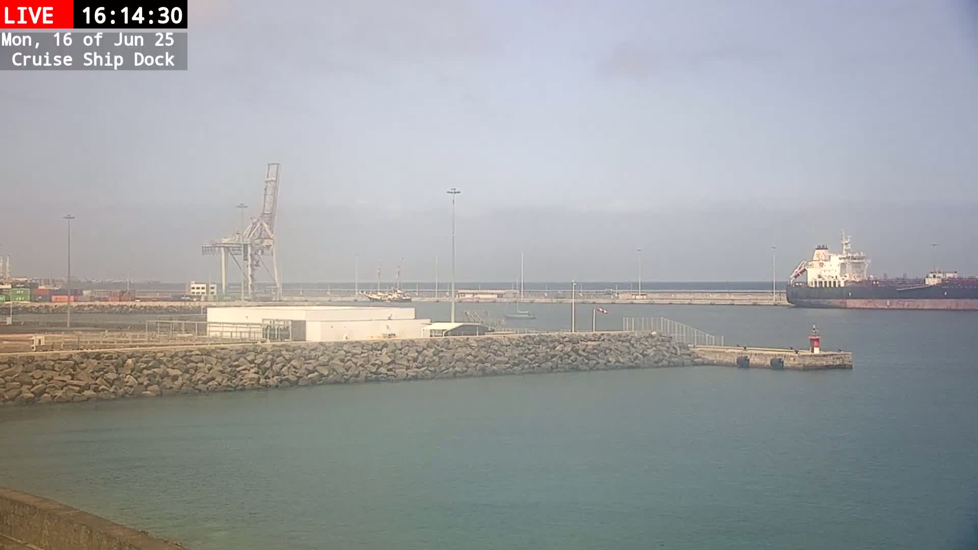 A hazy day at a cruise ship dock shows a large tanker in the harbor, a crane, and a rock wall separating the water from the dock.