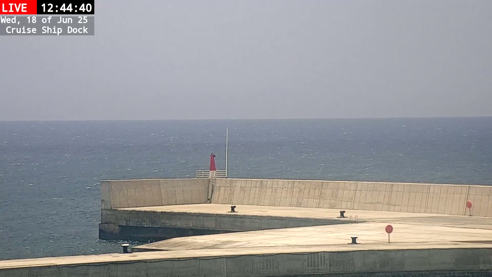 A concrete cruise ship dock extends into a calm, blue sea under a hazy sky.