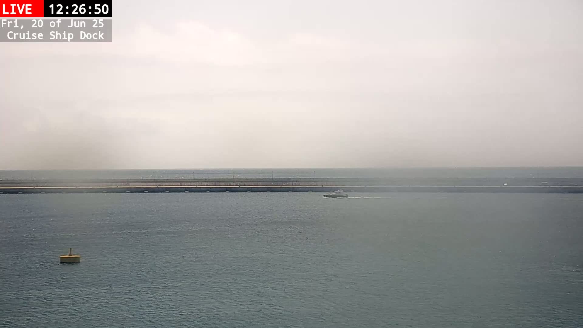 A hazy day shows a long pier extending into calm, gray water, with a small boat visible in the distance and a single buoy closer to the viewer.