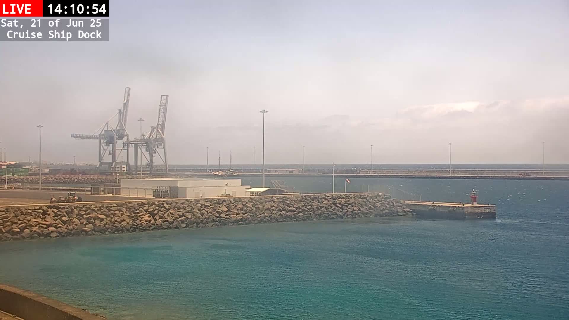 A hazy day reveals a cruise ship dock with several cranes, a stone wall, and a small lighthouse in the water.