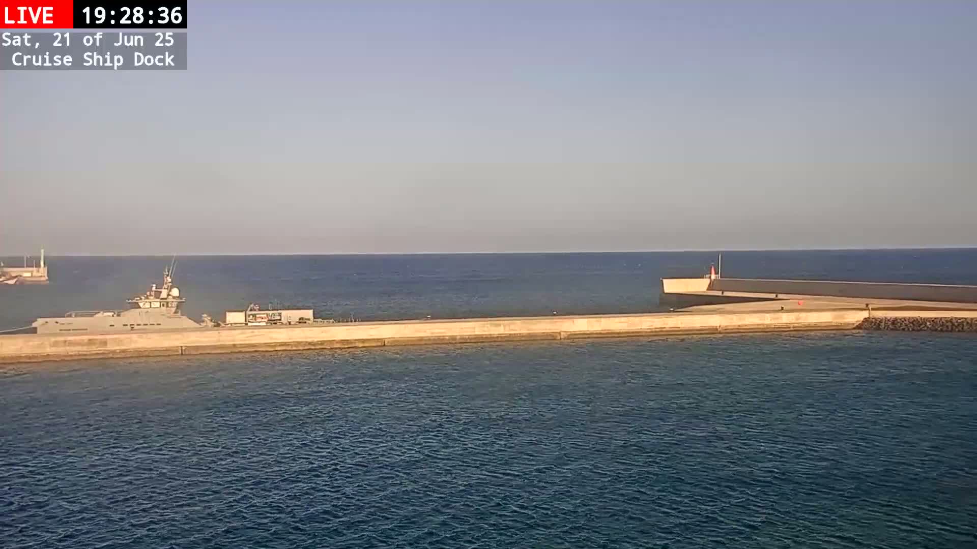 A long, gray ship is docked at a concrete pier extending into a calm, blue sea under a clear, light-blue sky.