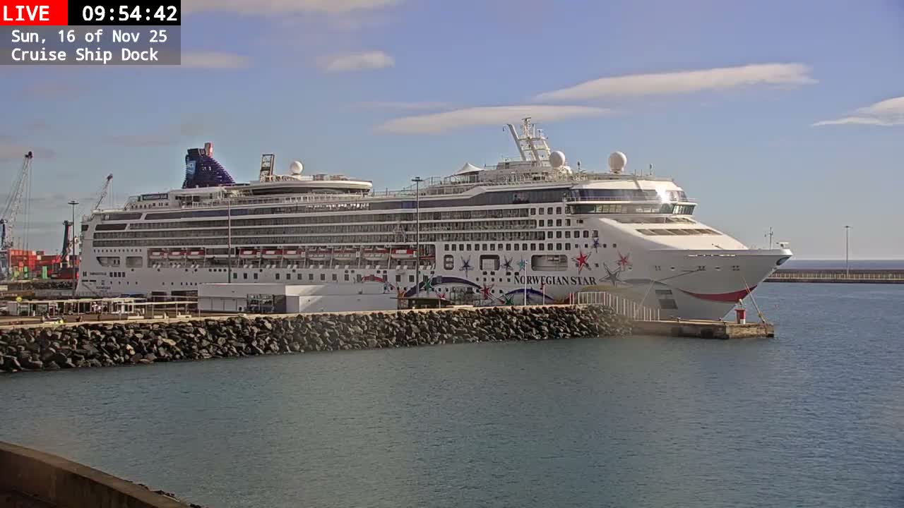 A large white cruise ship named "NORWEGIAN STAR" is docked at a pier with a rocky foreground and industrial port cranes in the background, under a clear and sunny sky with scattered clouds.