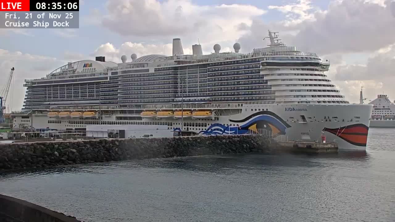 A large AIDAcosma cruise ship with a distinctive red and blue design is docked at a port, partially obscured by a rocky breakwater, under a partly cloudy sky, with another ship visible in the distance.