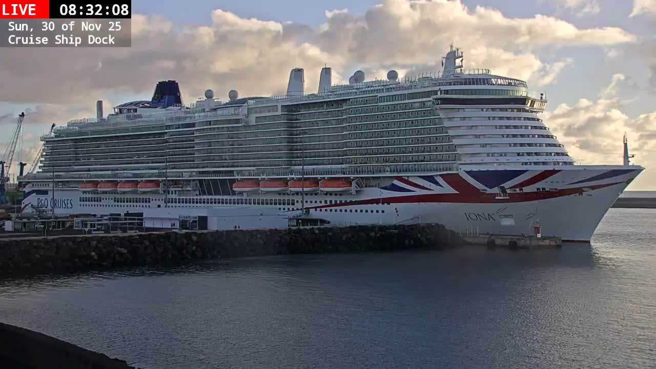 A large, multi-decked white cruise ship, featuring a prominent red, white, and blue design on its bow, is moored at a tranquil port, under a sky with scattered clouds.