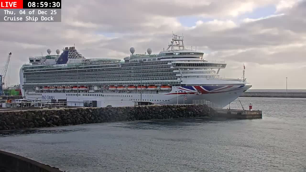 A large white cruise ship with red, white, and blue livery on its bow is docked in a port under a mostly cloudy sky, with calm water in the foreground and a distant breakwater.