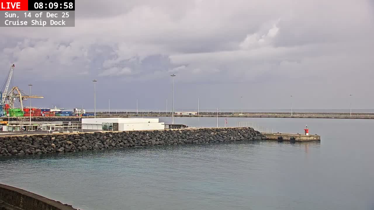 A port or cruise ship dock with a large crane, shipping containers, and buildings is seen under an overcast sky, with a rocky breakwater extending into calm grey water towards a small red and white lighthouse beacon.