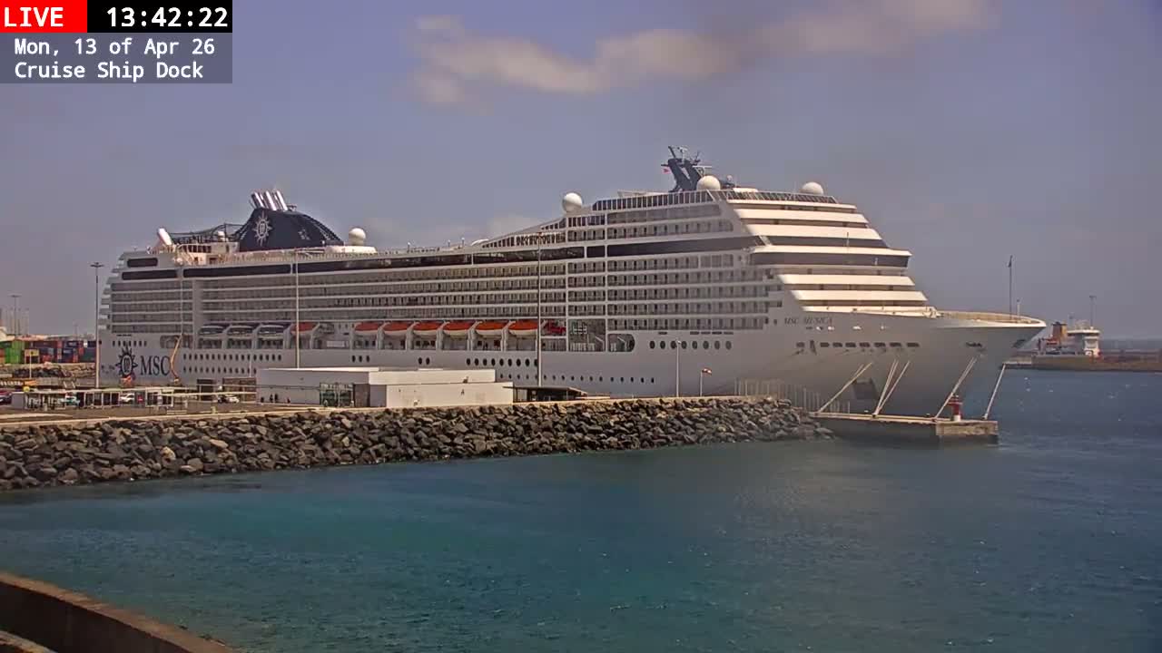 A port or cruise ship dock with a large crane, shipping containers, and buildings is seen under an overcast sky, with a rocky breakwater extending into calm grey water towards a small red and white lighthouse beacon.