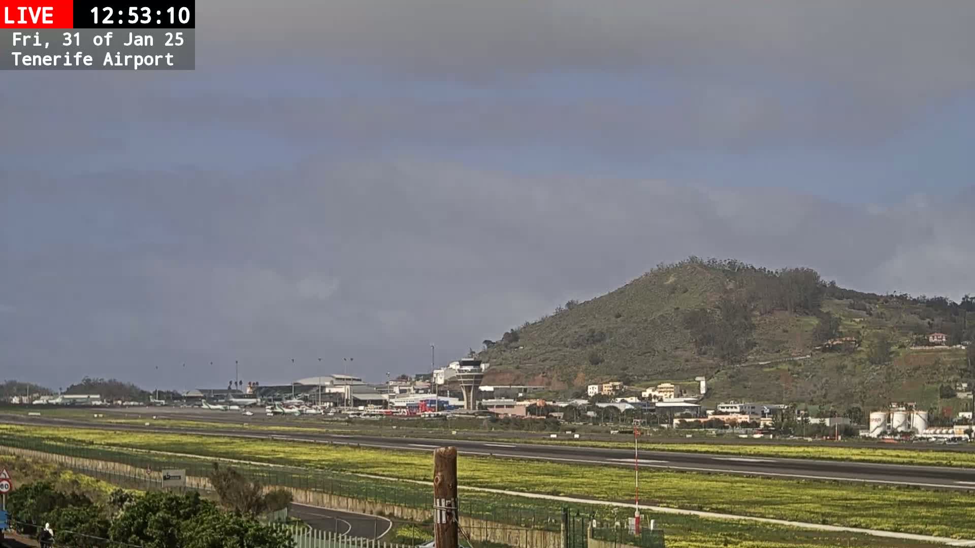 An airport runway with several airplanes is visible, situated below a gently sloping, green hill under a mostly cloudy sky.